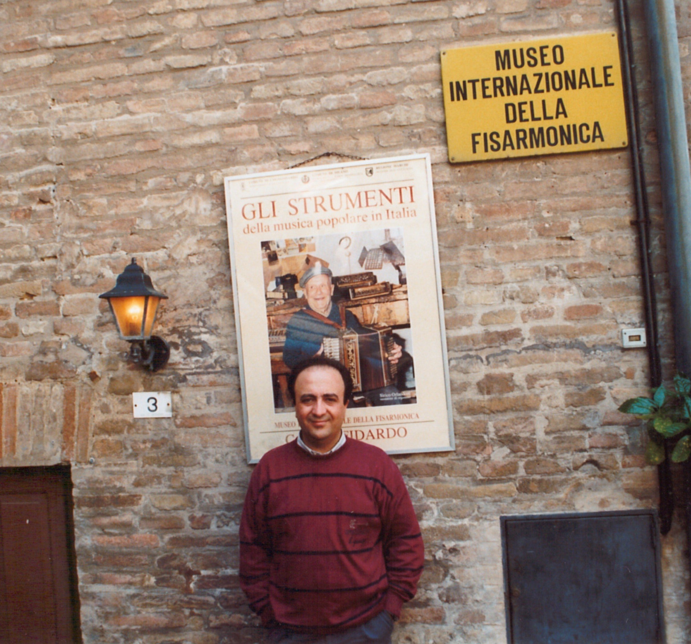Uomo con maglione a righe rosse e nere che si trova davanti a un muro di mattoni, con poster e luci sulla parete.