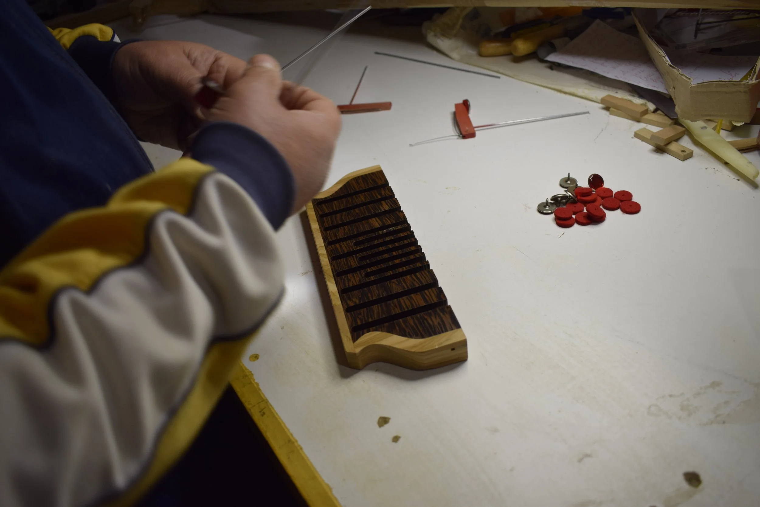 Una persona sta lavorando su un tavolo con vari strumenti e materiali, include un pezzo di legno usato come supporto o decorazione, bottoni e altri oggetti di bricolage.