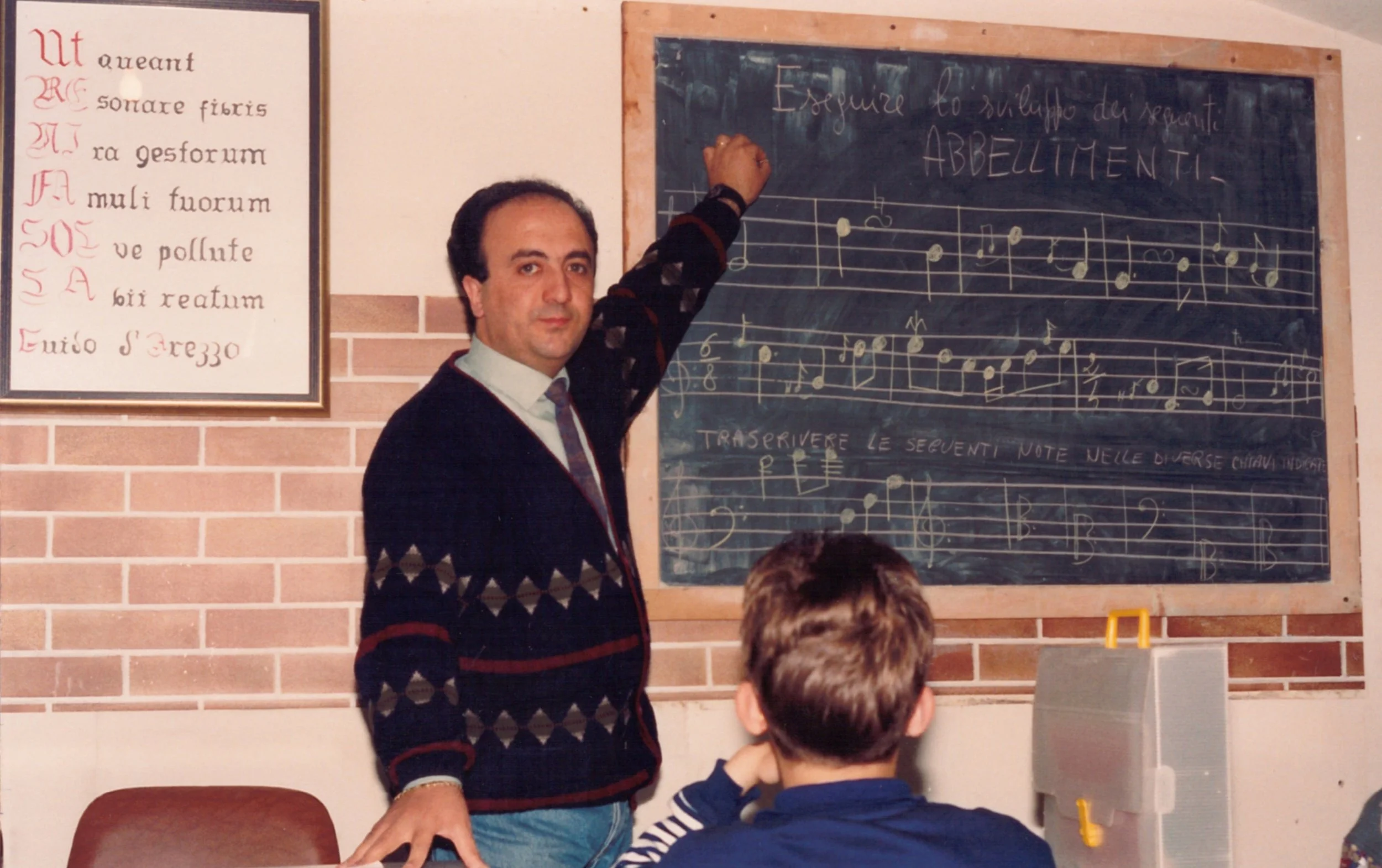 Un insegnante maschio di fronte a una lavagna di scuola, scrive partiture musicali. In piedi, indossa un maglione con un motivo a rombi, camicia bianca e cravatta, mentre gli studenti seduti osservano. La stanza ha pareti di mattoni, e nel lato sinistro c'è un cartellone con una poesia in italiano.