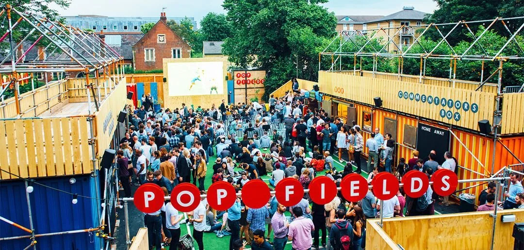A lively outdoor event at Pop Fields with many people gathered in front of a large outdoor screen, surrounded by colourful wooden structures and seating areas, under a bright sky. Pop Brixton.