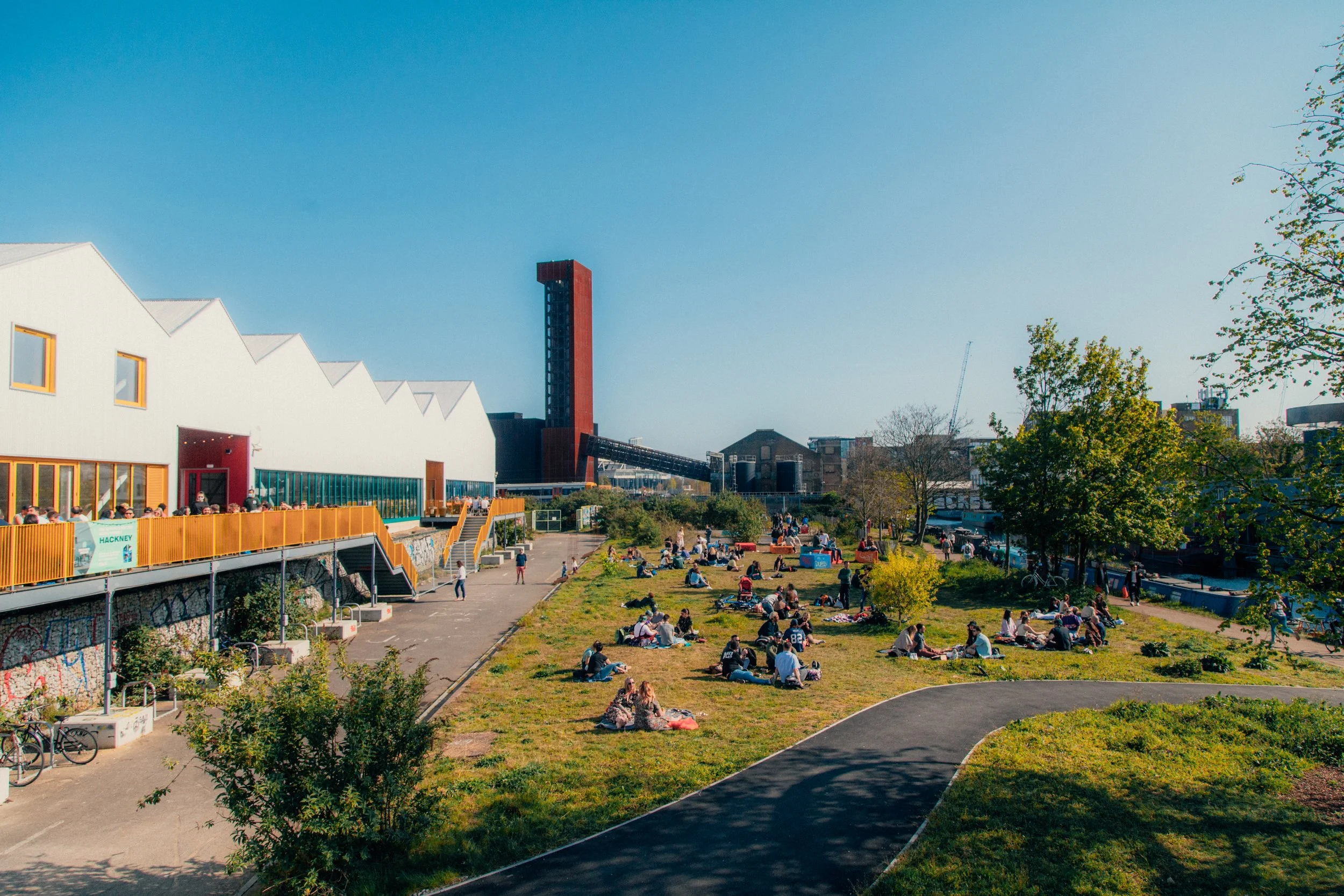 A park filled with people sitting on the grass and walking, with a modern white building with orange windows and a tall red tower in the background, under a clear blue sky. Hackney Bridge.