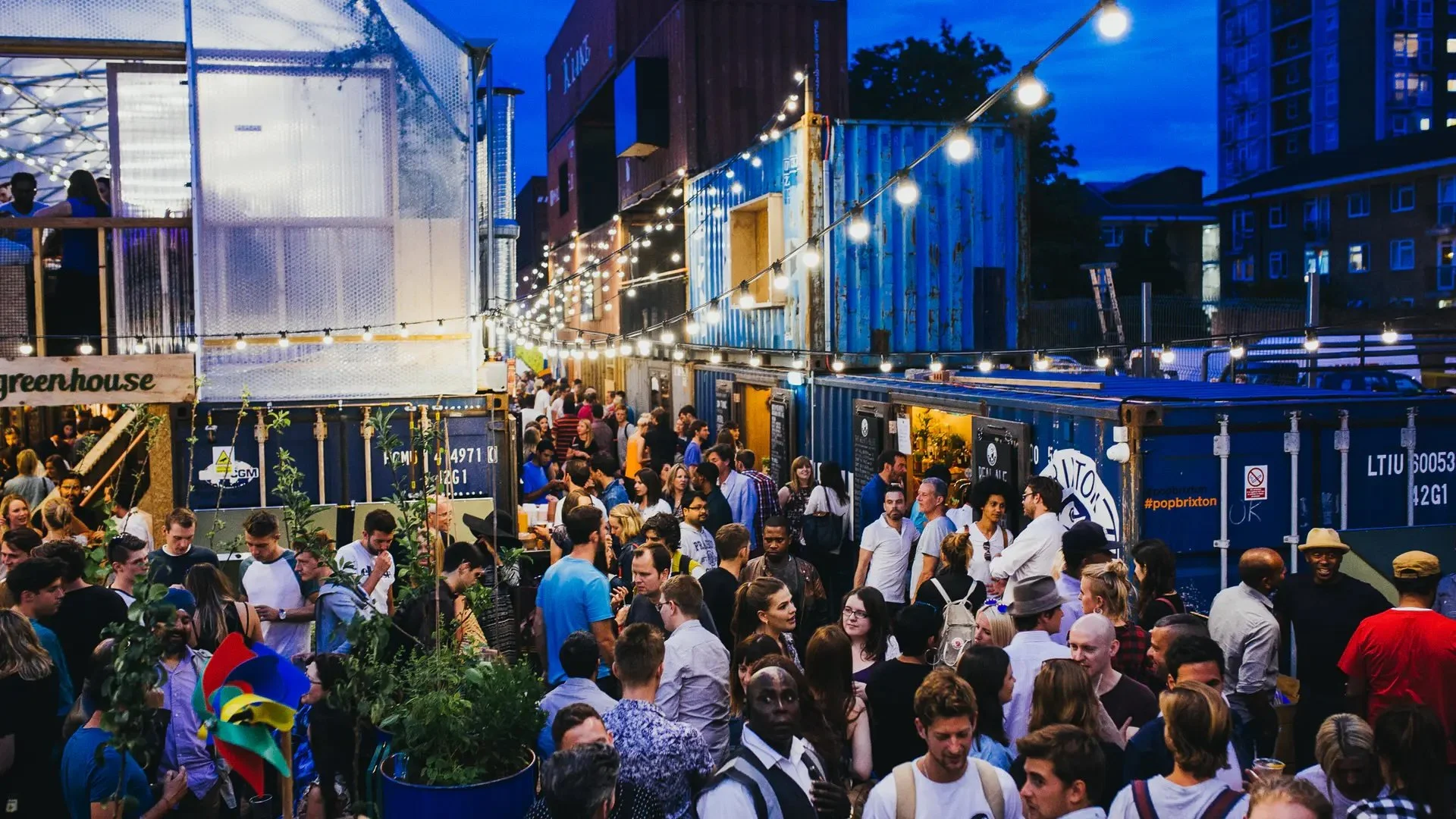Crowd of people at an outdoor evening event with string lights overhead, inside a space made of repurposed shipping containers, some with illuminated signs. Pop Brixton.