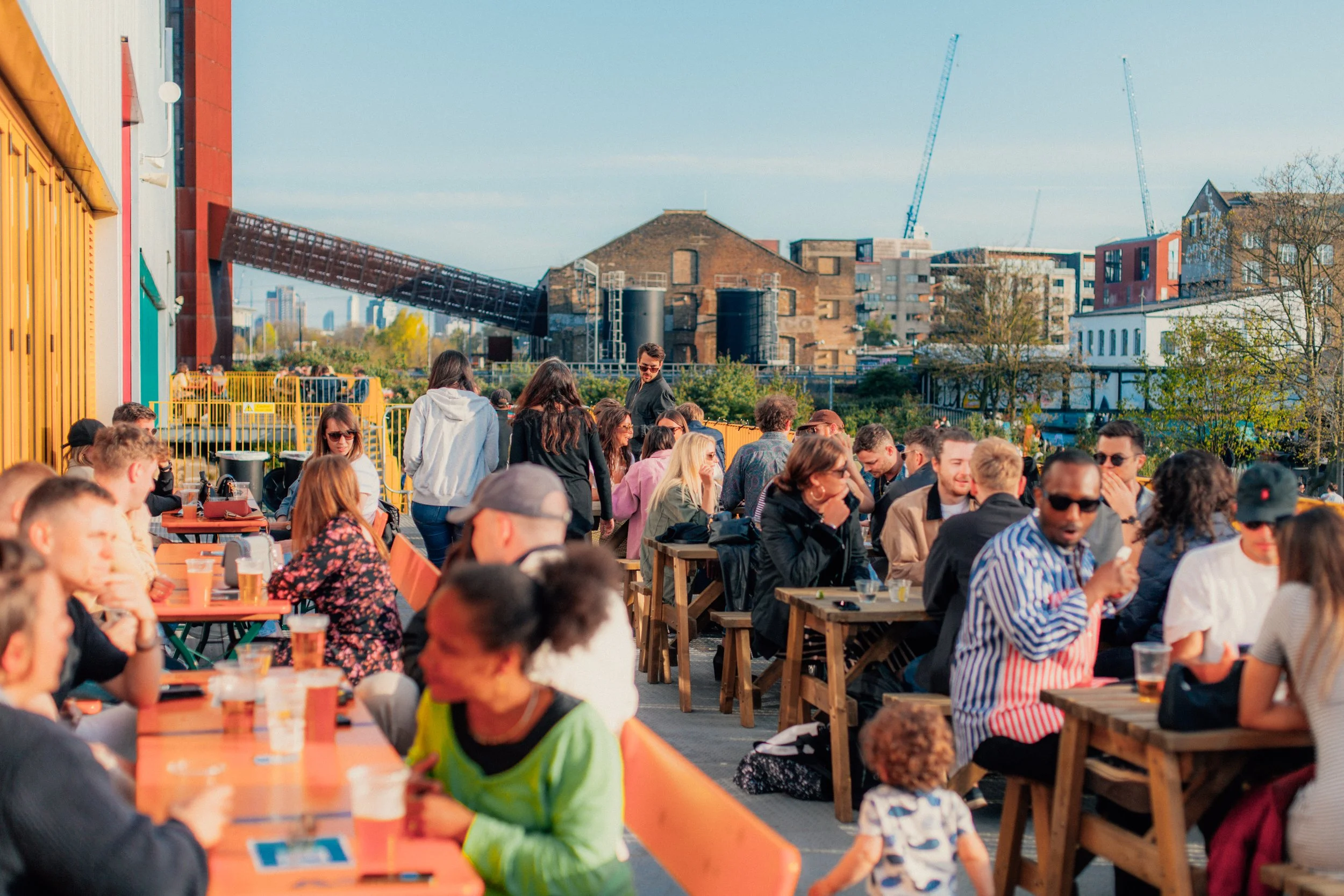 A diverse group of people sitting and socialising at outdoor tables in an urban setting with buildings and construction cranes in the background. Hackney Bridge.