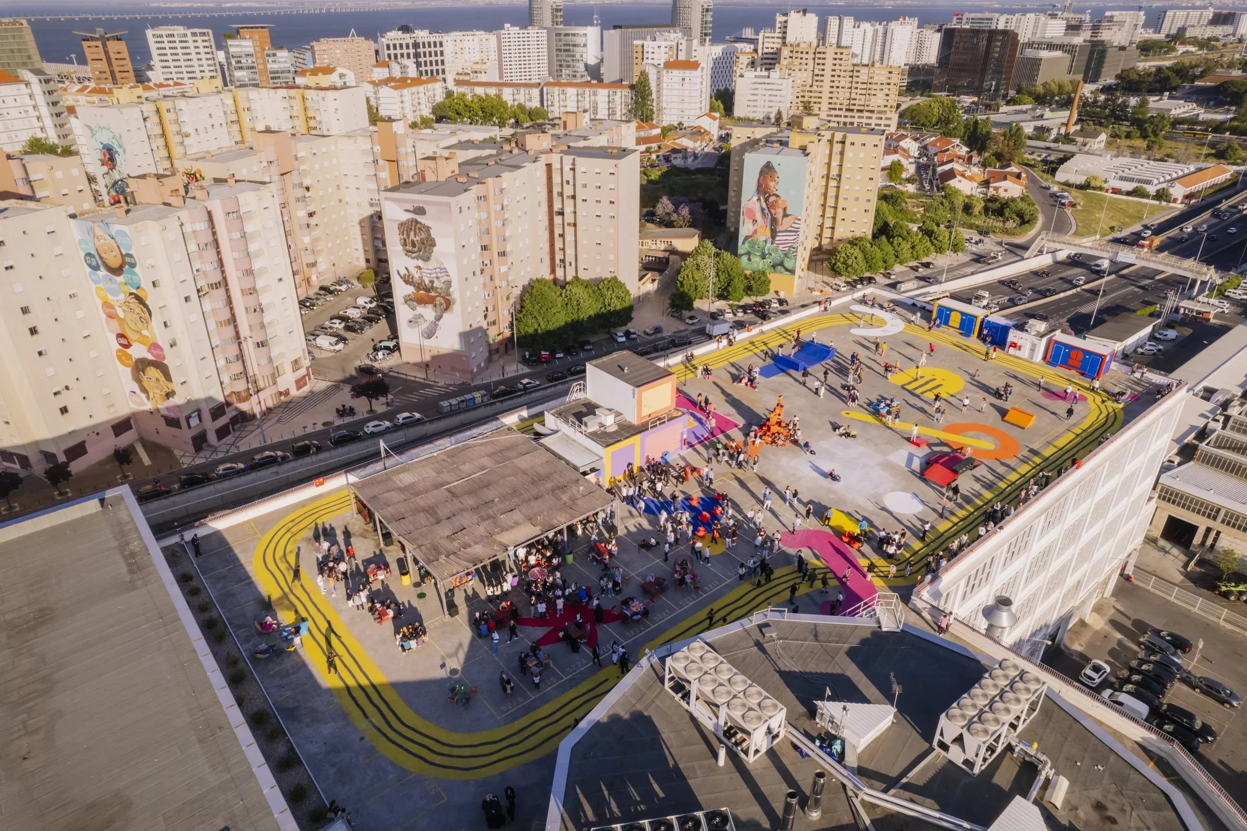 Aerial view of a colorful outdoor playground with children and adults, surrounded by high-rise buildings and murals on apartment walls, in a cityscape. IDB Lisbon.