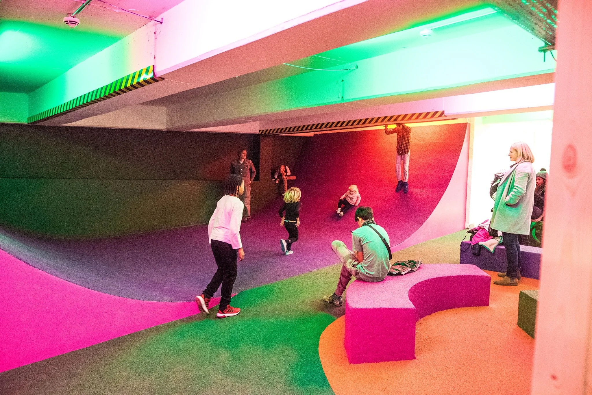 Indoor playground with colourful ramps and walls, children and adults playing and sitting, illuminated with pink, green, and orange lights. Peckham Levels.