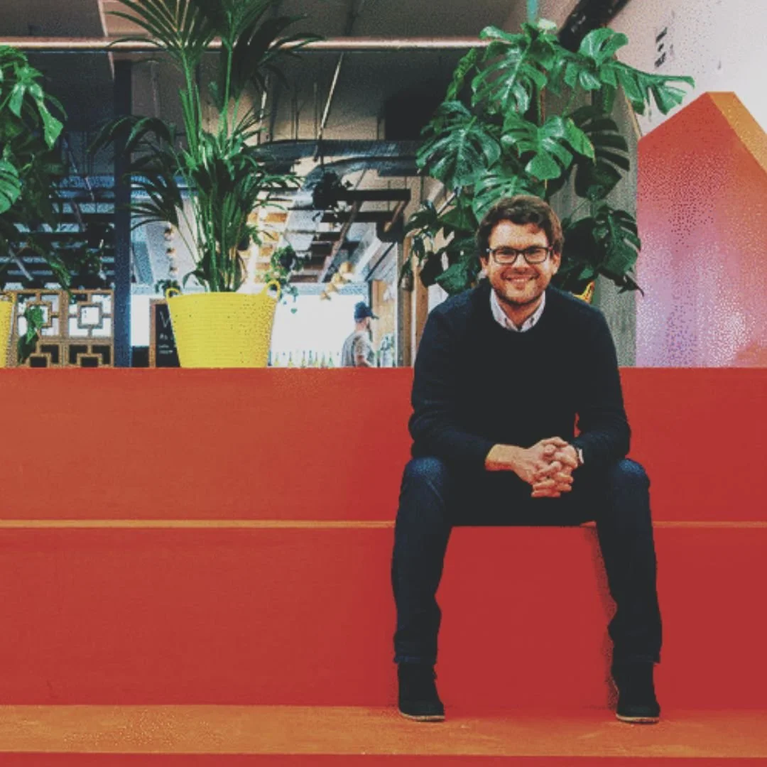 A man with glasses, wearing a black sweater and jeans, sitting on a red bench in an indoor space with large green plants in yellow pots behind him, and a person walking in the background. James Leay. Peckham Levels.