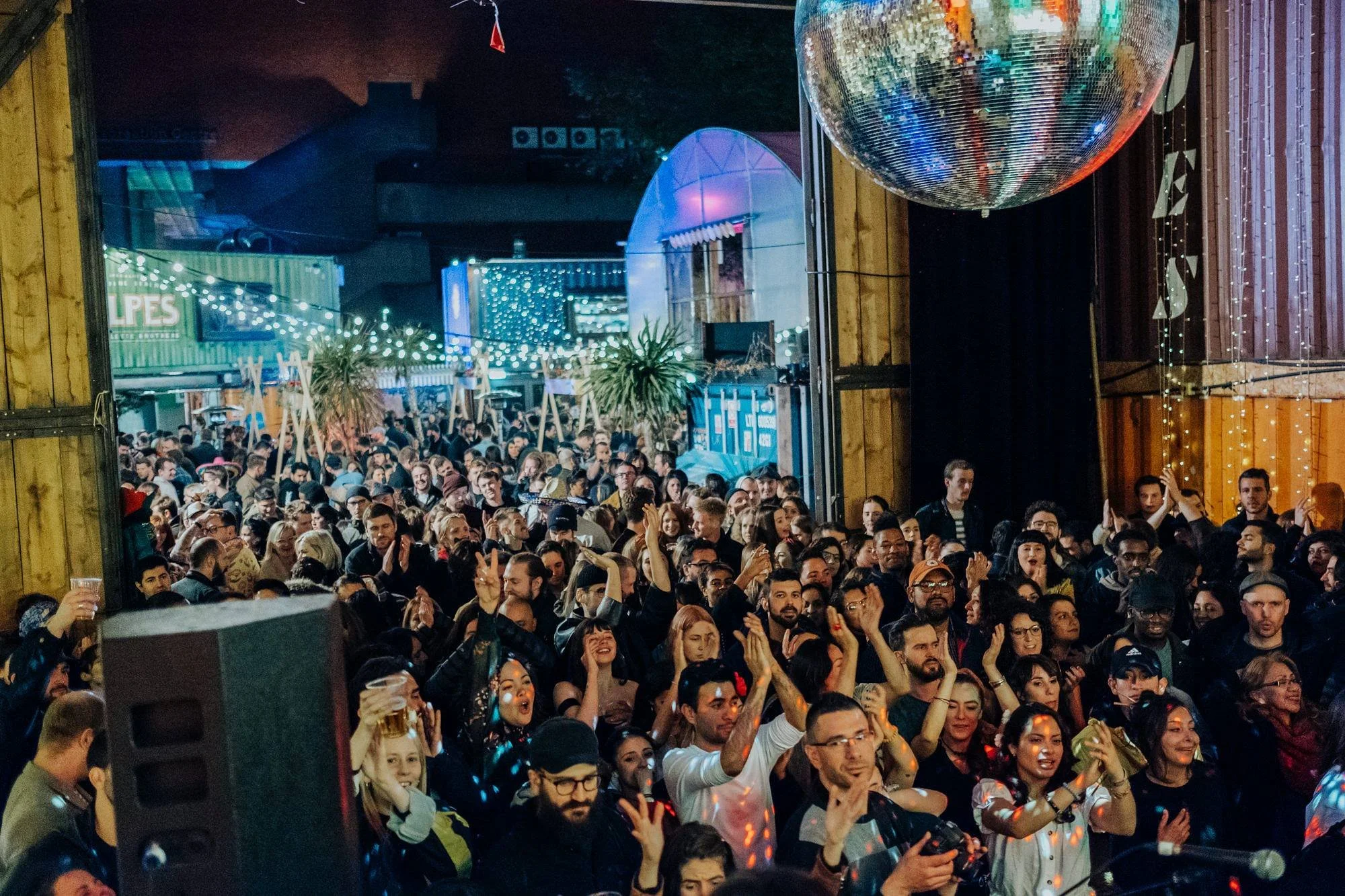 A large crowd of people at an indoor concert or party, some with drinks, enjoying and dancing with colorful lighting and a disco ball overhead. Pop Brixton.