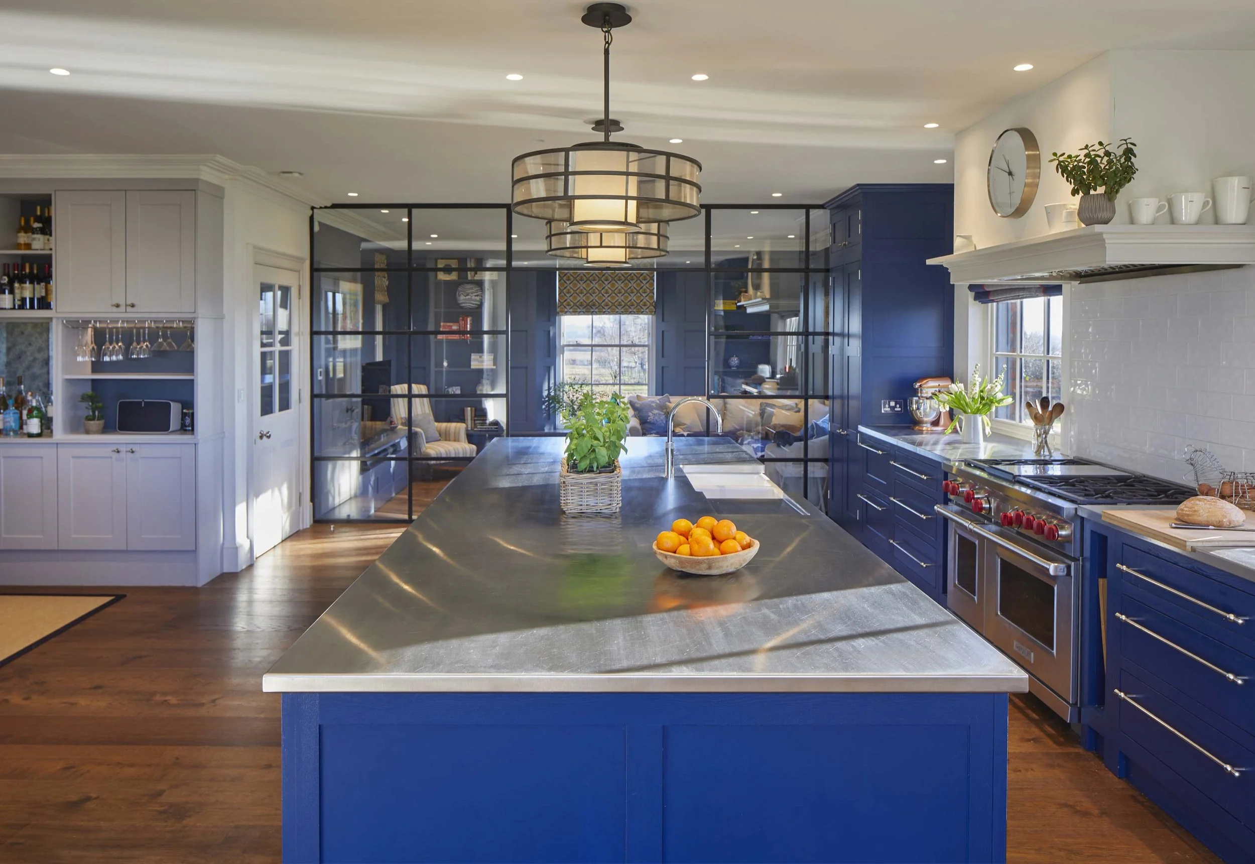 A modern kitchen with a large blue island, gray cabinets, and a stone countertop. The kitchen includes a bowl of oranges, potted herbs, and various kitchen appliances. There's a black-framed glass door separating the kitchen from a cozy sitting area with furniture and a window letting in natural light.