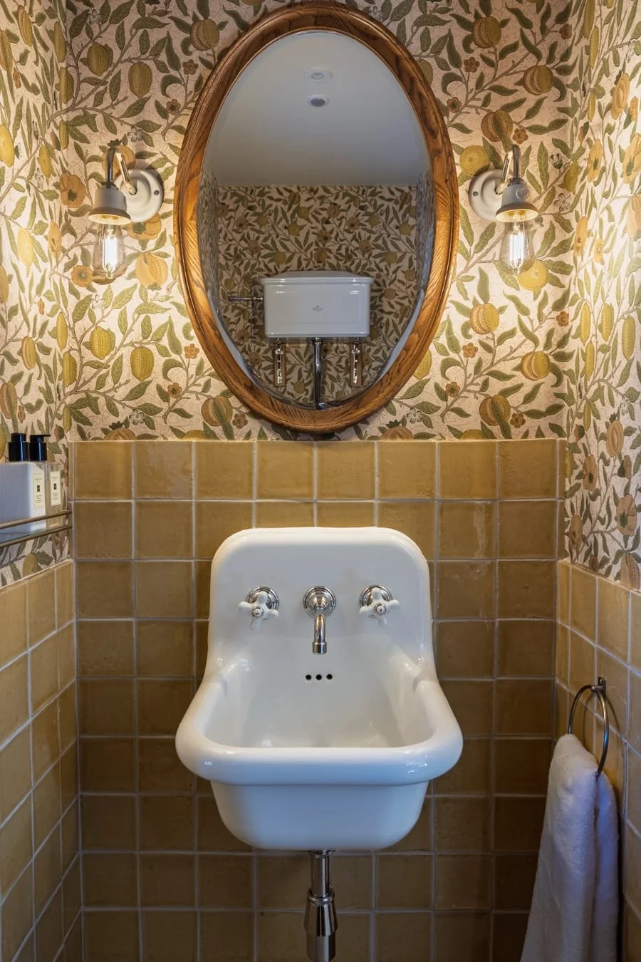 A vintage-inspired bathroom with a wall-mounted sink, decorated with floral wallpaper, a large oval wooden framed mirror, wall sconces with Edison bulbs, yellow tiles, and a towel ring with a white towel.