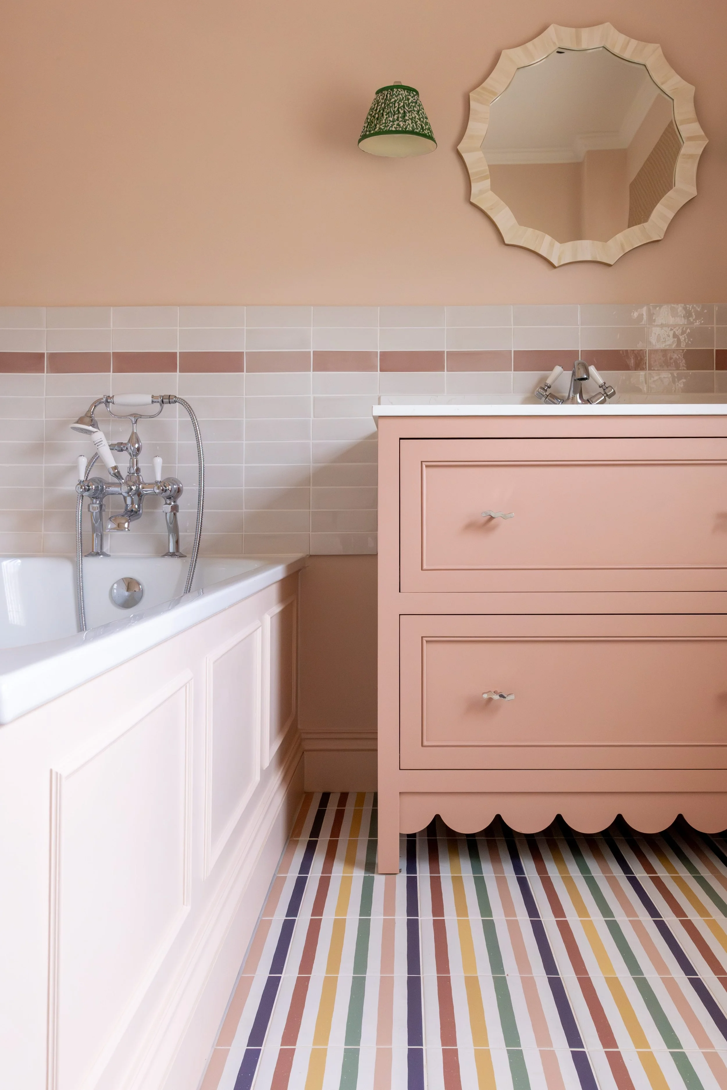 Bathroom with pink walls, a pink cabinet, a mirror with a scalloped wooden frame, striped multicolored floor tiles, a white bathtub, and a wall-mounted green lamp.