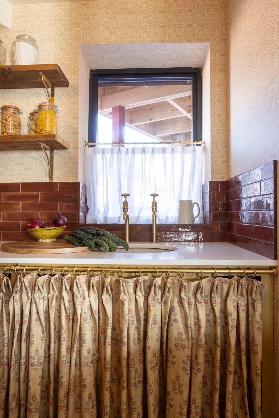Kitchen with a window above the sink, gold faucet, red brick backsplash, patterned curtain, and shelves with jars and food items.