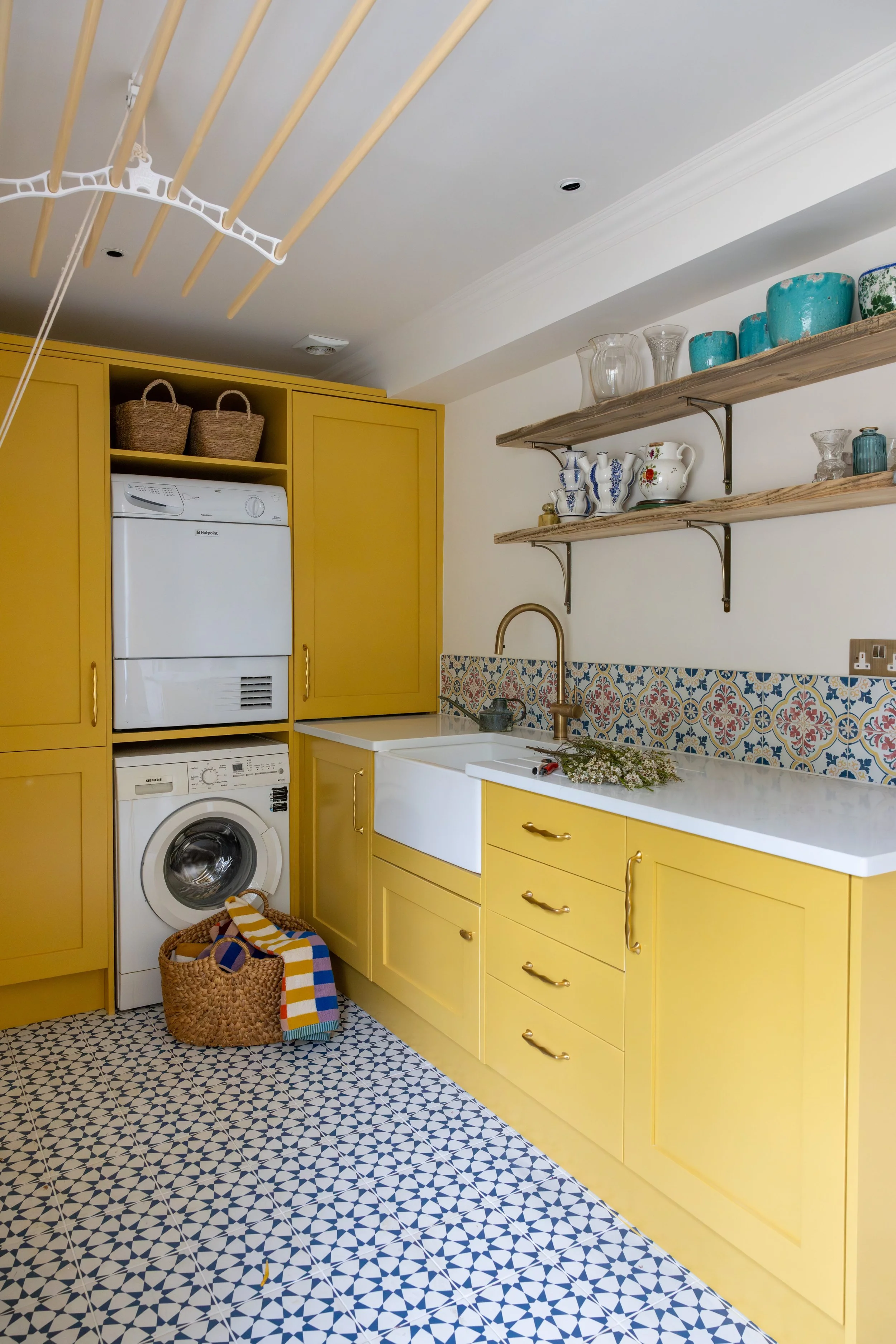A yellow kitchen with a white sink, patterned tile backsplash, open shelves with dishes, and a laundry area with washing and drying machines stacked. Basket with colorful striped towels on the floor.