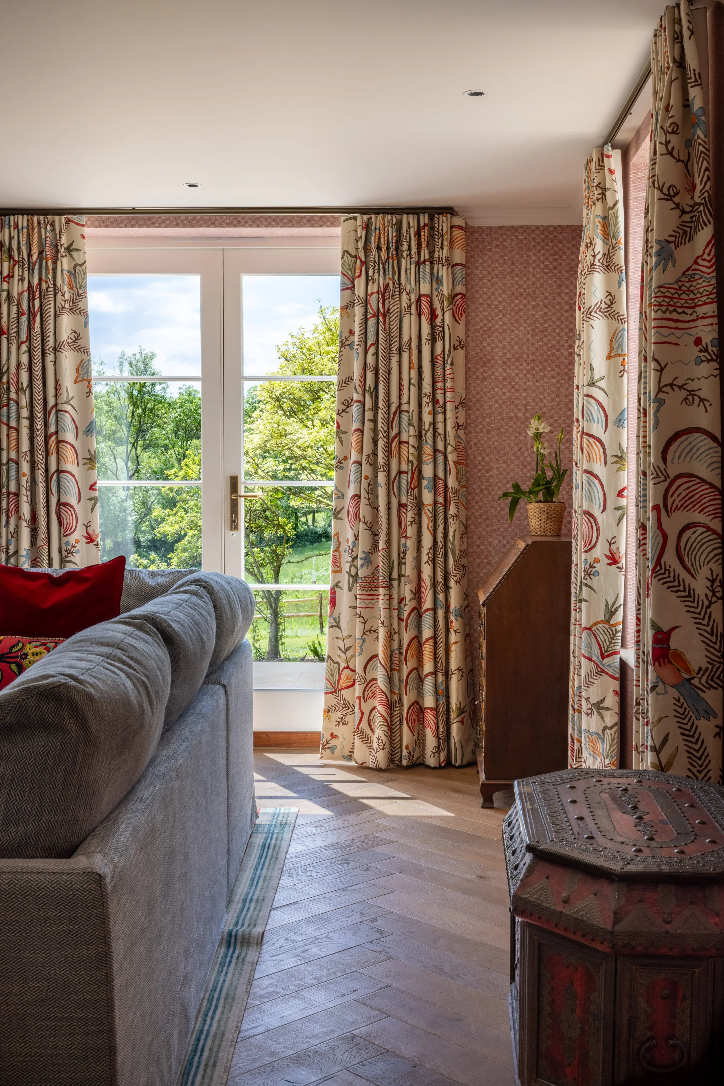 Living room with large window and view of green trees outside, patterned curtains, a gray sofa with a red pillow, wooden floor, and an antique wooden cabinet with a potted plant on top.
