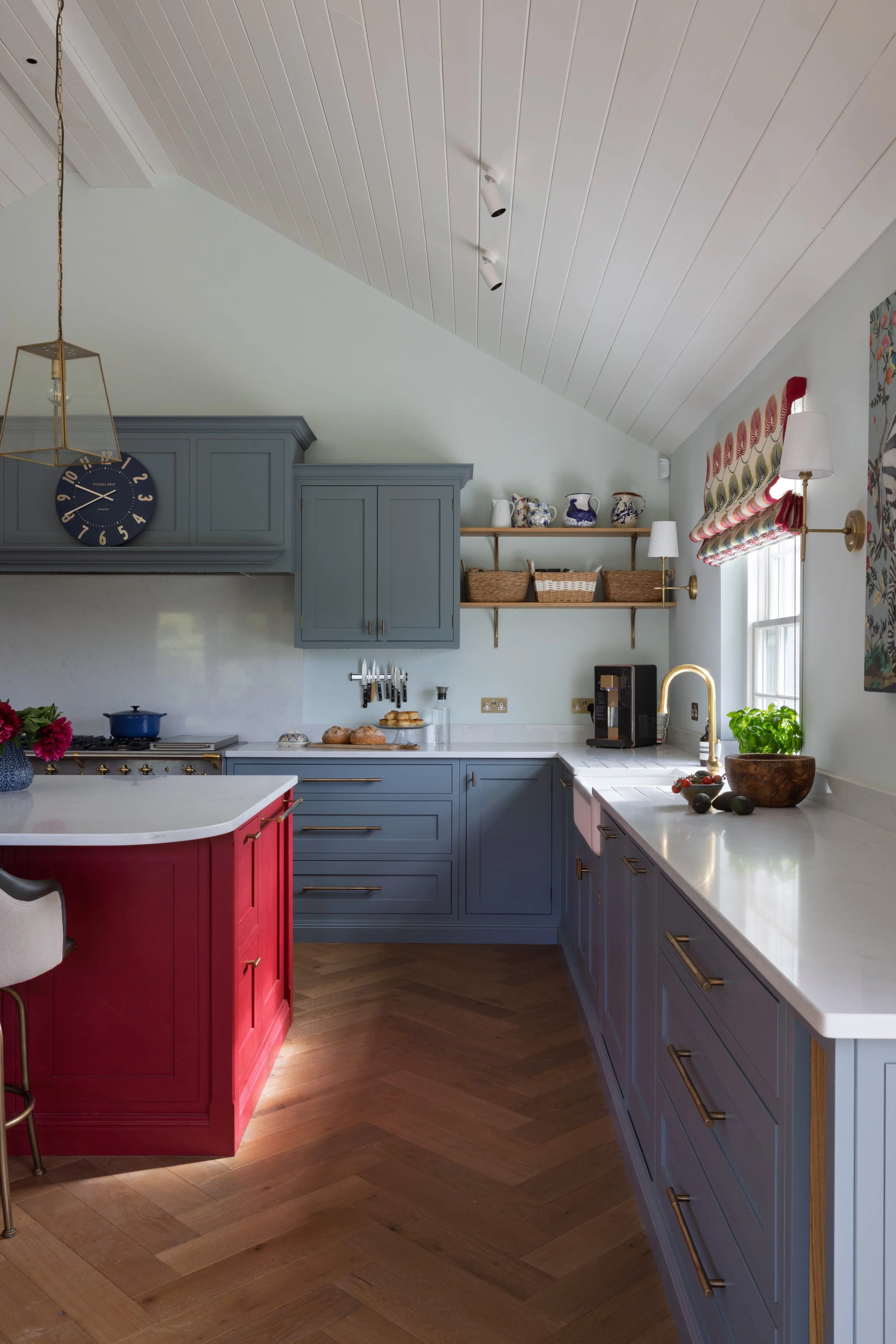 A modern kitchen with blue and red cabinetry, white countertops, hanging shelves with decorative vases, a window with colorful curtains, and wooden flooring.