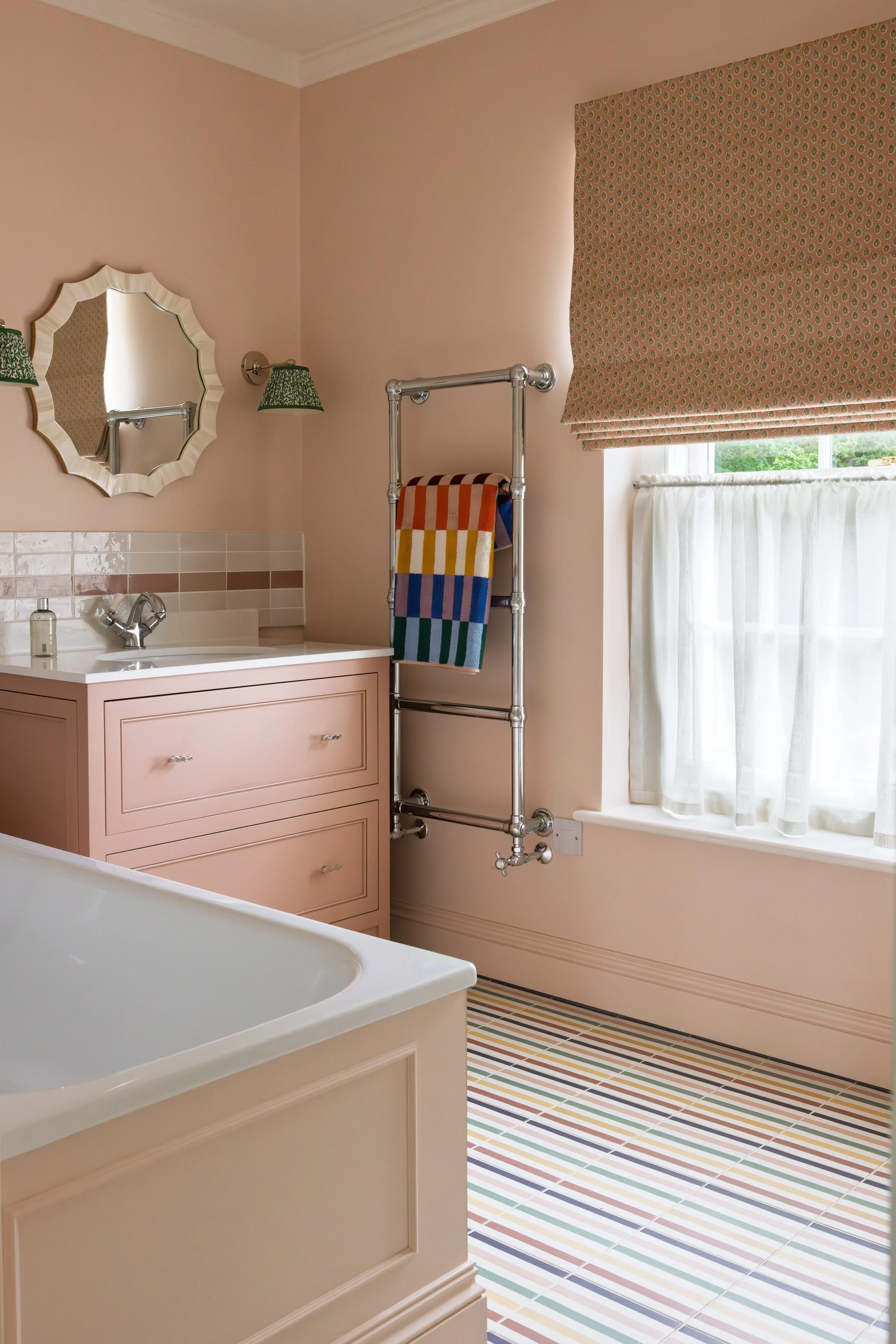 A bathroom featuring a pink vanity with drawers, a striped multicolored bath mat on the floor, a bathtub, a wall-mounted mirror, green lampshades, a window with a patterned Roman shade, a towel rack with a colorful towel, and striped floor tiles.