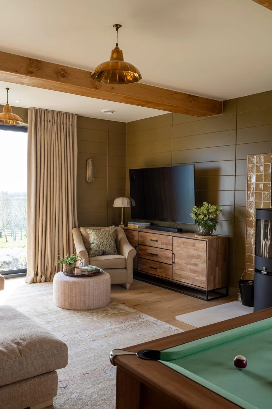 A cozy living room with beige armchair, wooden TV stand, large flat-screen TV, beige curtains, potted plant, table lamp, pool table, wood beam ceiling, warm lighting, and scenic view outside window.