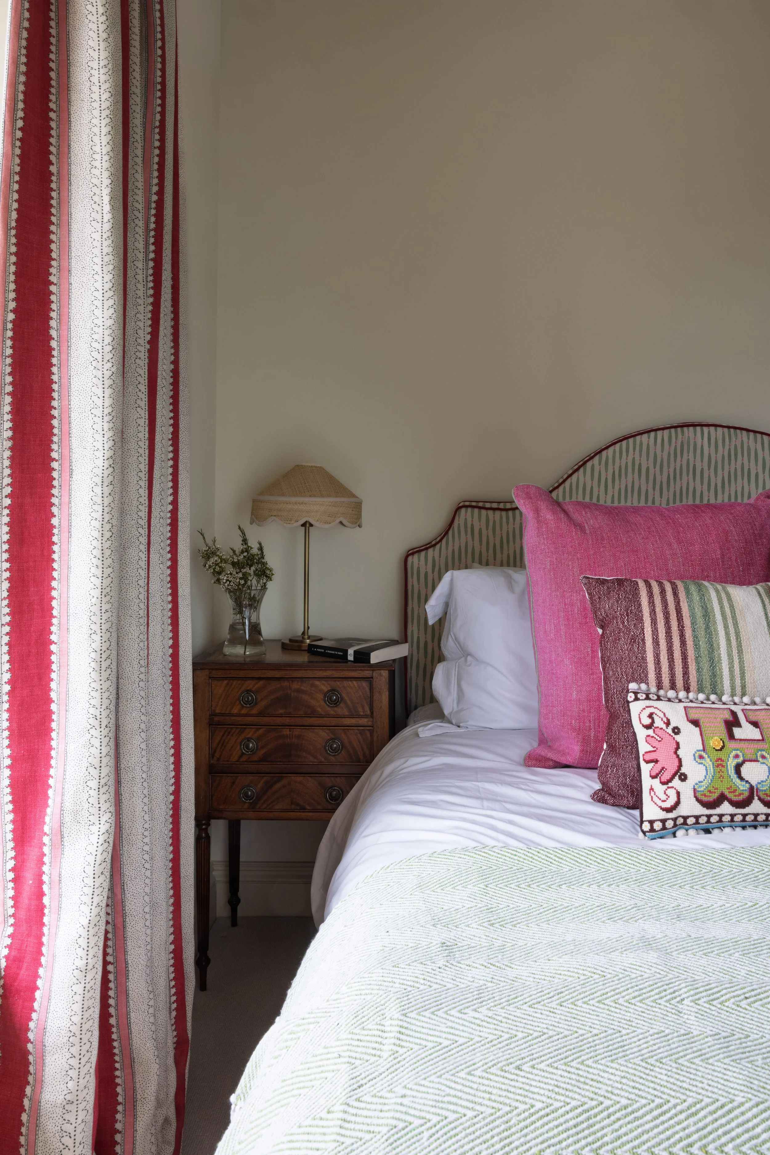 A bedroom with a bed that has white bedding, decorative pillows, and a wooden nightstand with a lamp, a book, and a small plant. There are red and white curtains on the window.