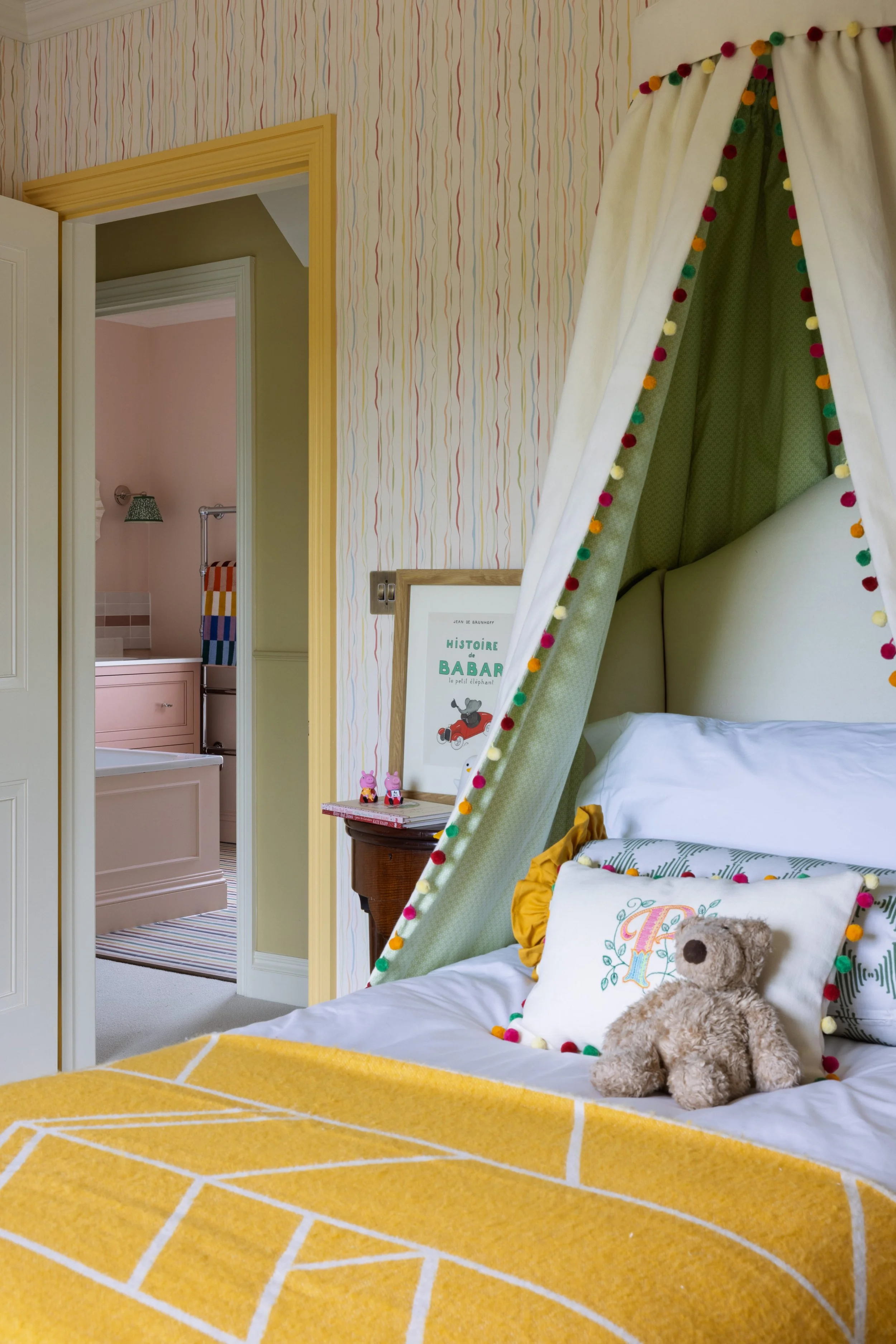 A bedroom with a bed that has a pillow and a teddy bear. Yellow and white bed linens, a canopy with colorful trim, striped wallpaper, and a yellow rug. Visible doorway leading to a bathroom with pink walls and striped carpet.