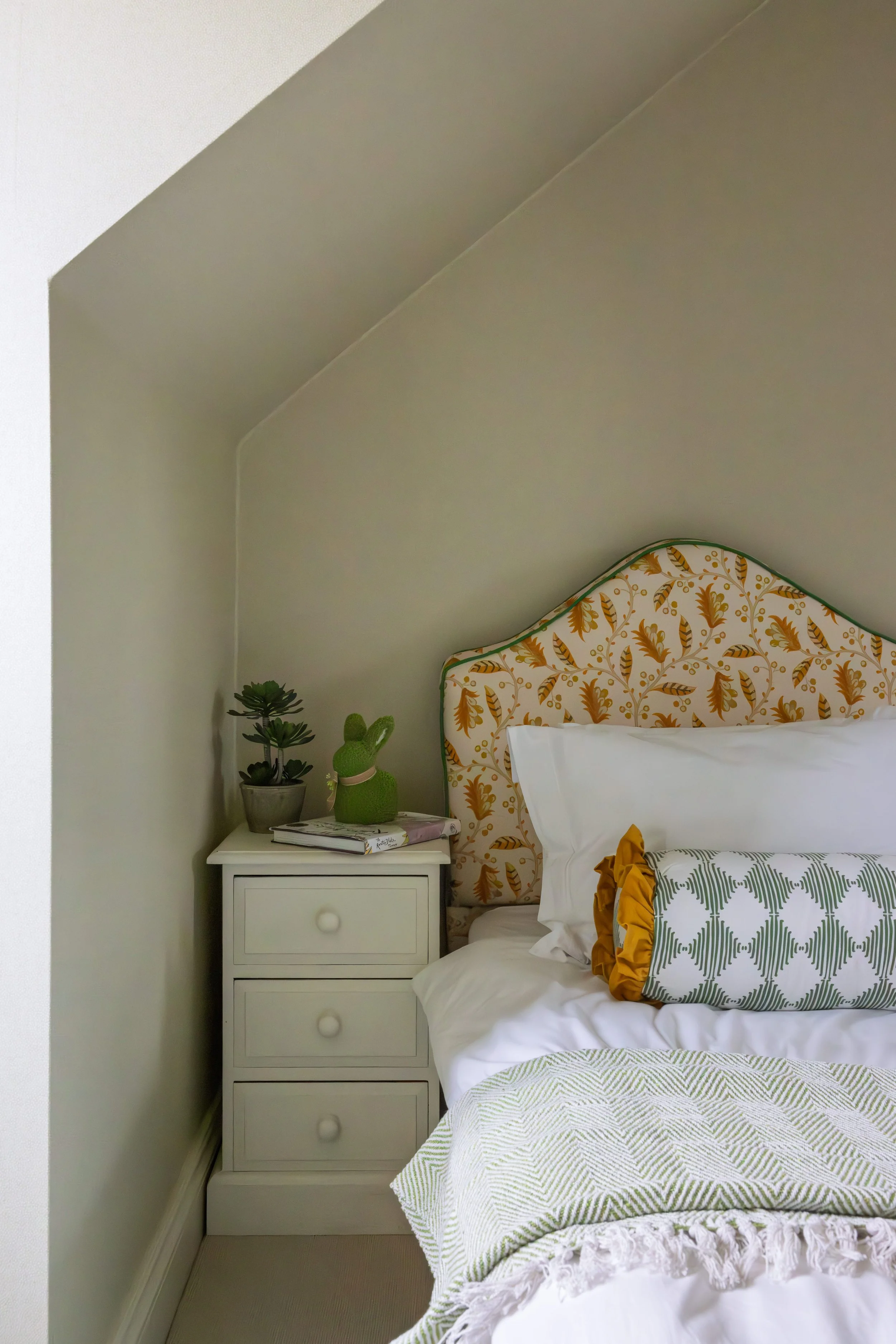 Close-up of a bedroom with a white nightstand, green plants, decorative pillows, and a bed with a patterned headboard and linens.