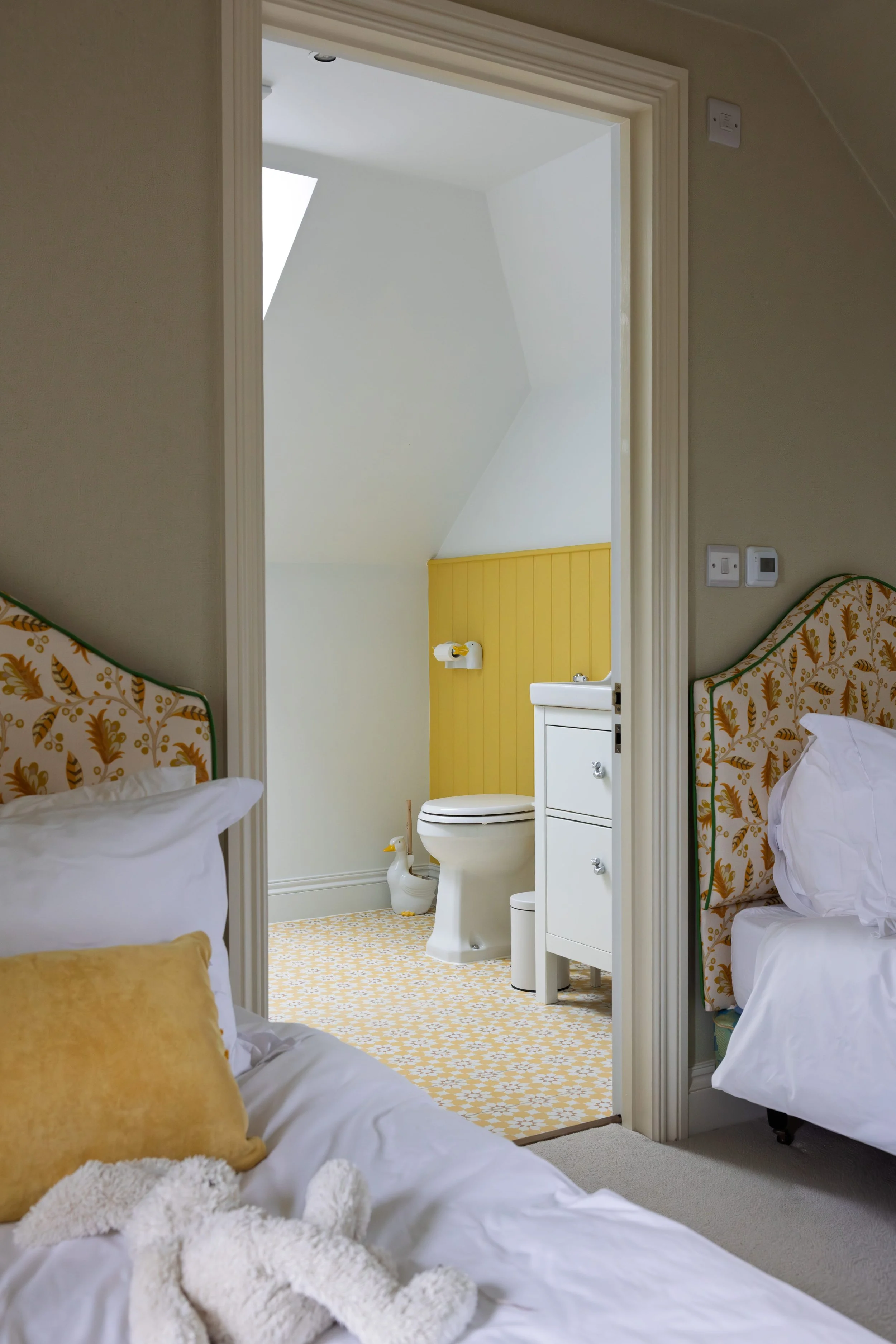 A view from two beds in a bedroom looking into an adjacent bathroom with a white toilet, a small white cabinet, and a duck-shaped trash bin. The bathroom has yellow paneling on the wall and a patterned yellow and white tile floor. The bedroom has bei