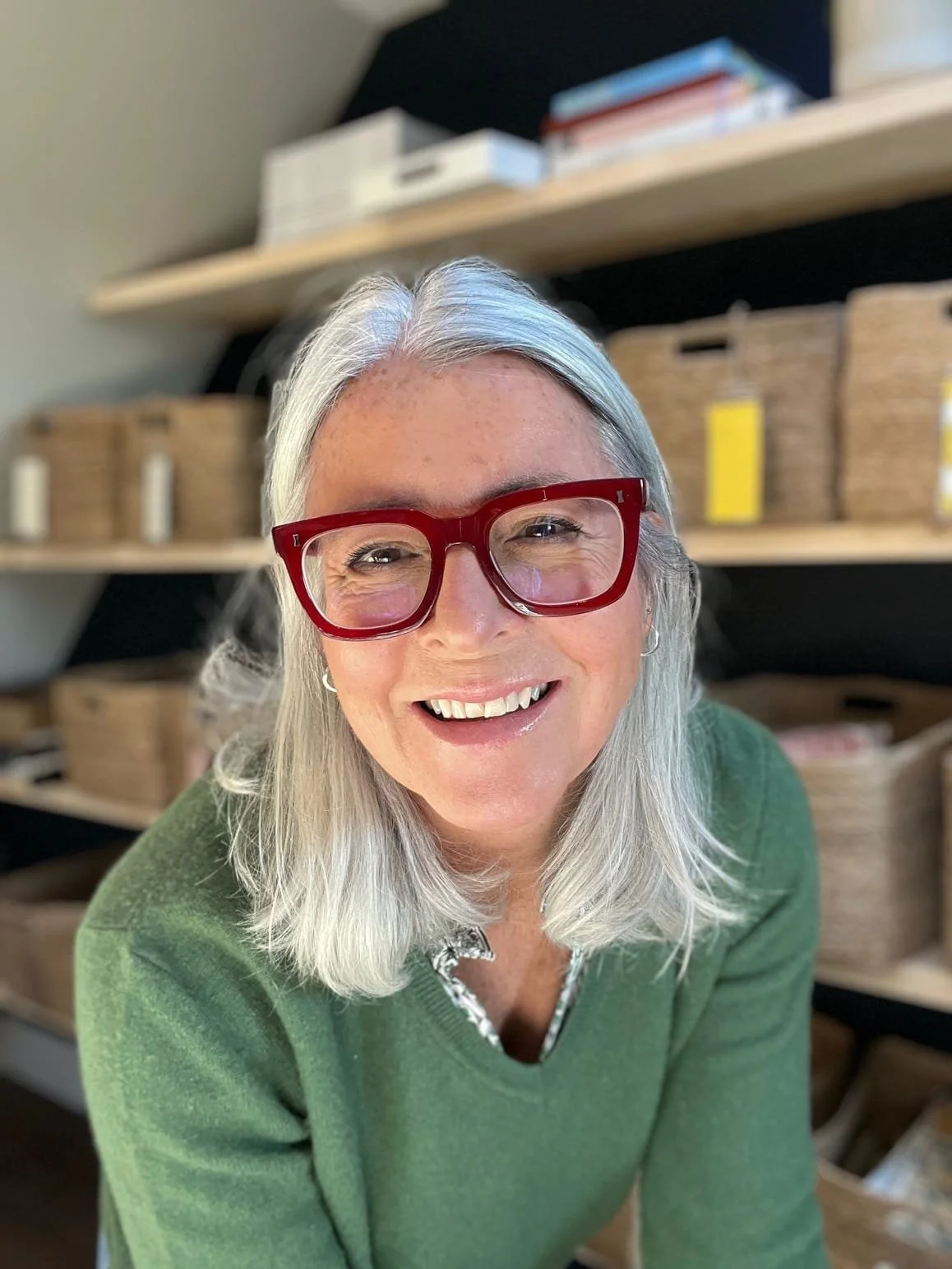 A smiling woman with gray hair wearing red glasses and a green sweater, standing in front of shelves with baskets and files.