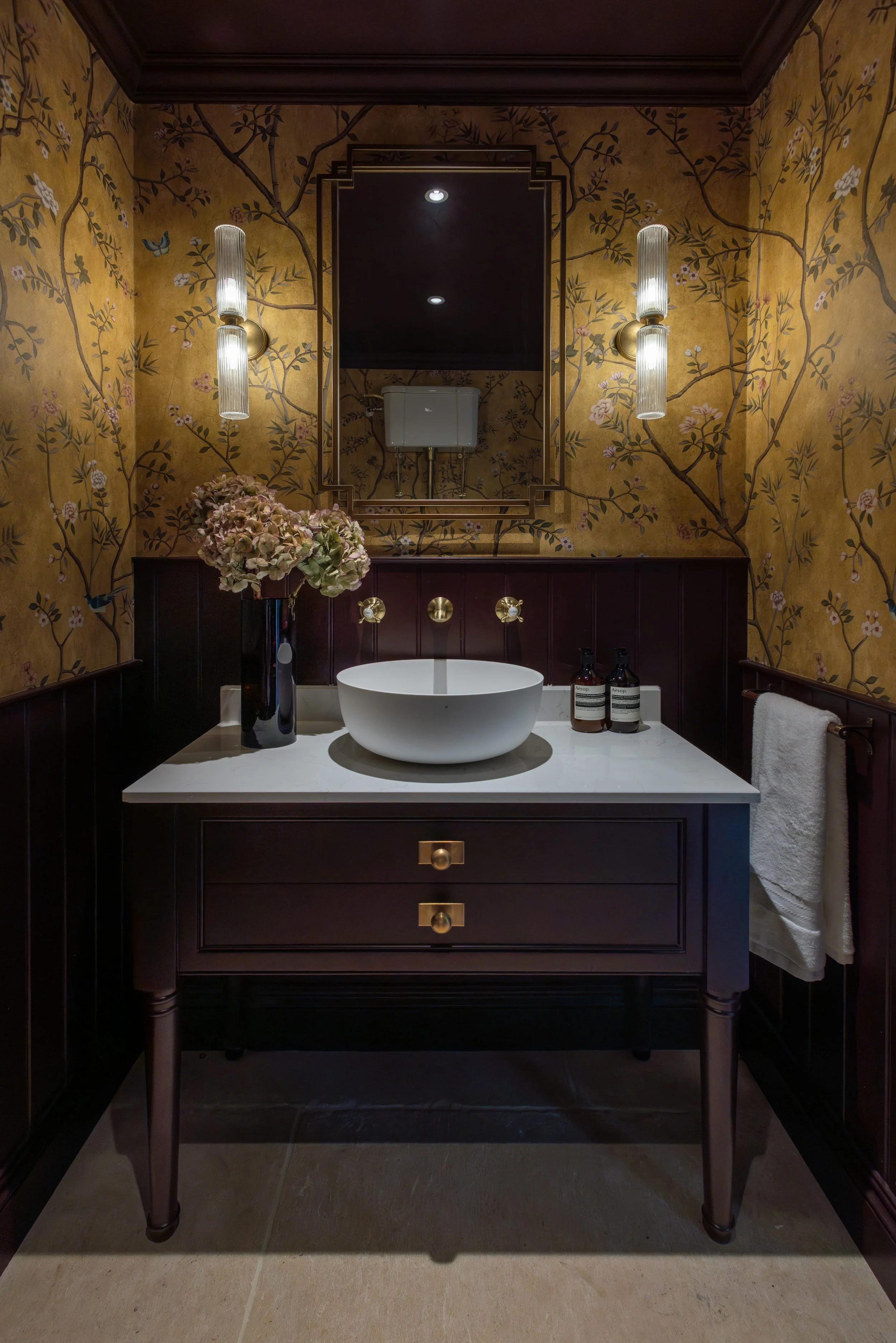 A bathroom sink with a white vessel basin on a dark wood vanity with three drawers featuring gold knobs. Behind the sink, a large vase with pinkish flowers is placed. Two wall-mounted light fixtures with ribbed glass shades are on either side of a fr