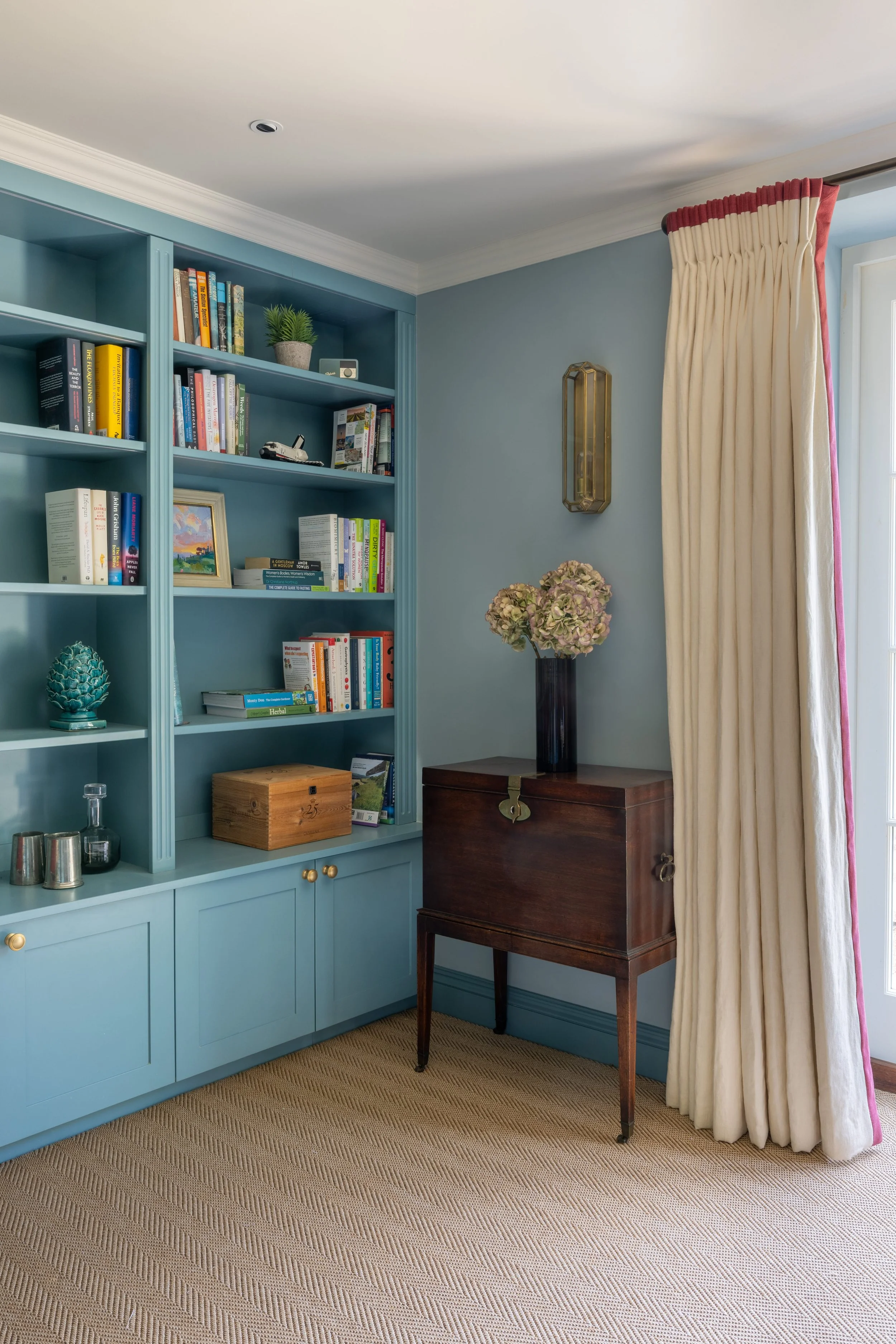 A living room corner with a blue built-in bookshelf filled with books and decorative items, a dark wooden cabinet with pink hydrangeas in a black vase on top, beige curtains partially covering a window, a gold wall sconce, and a beige textured carpet.