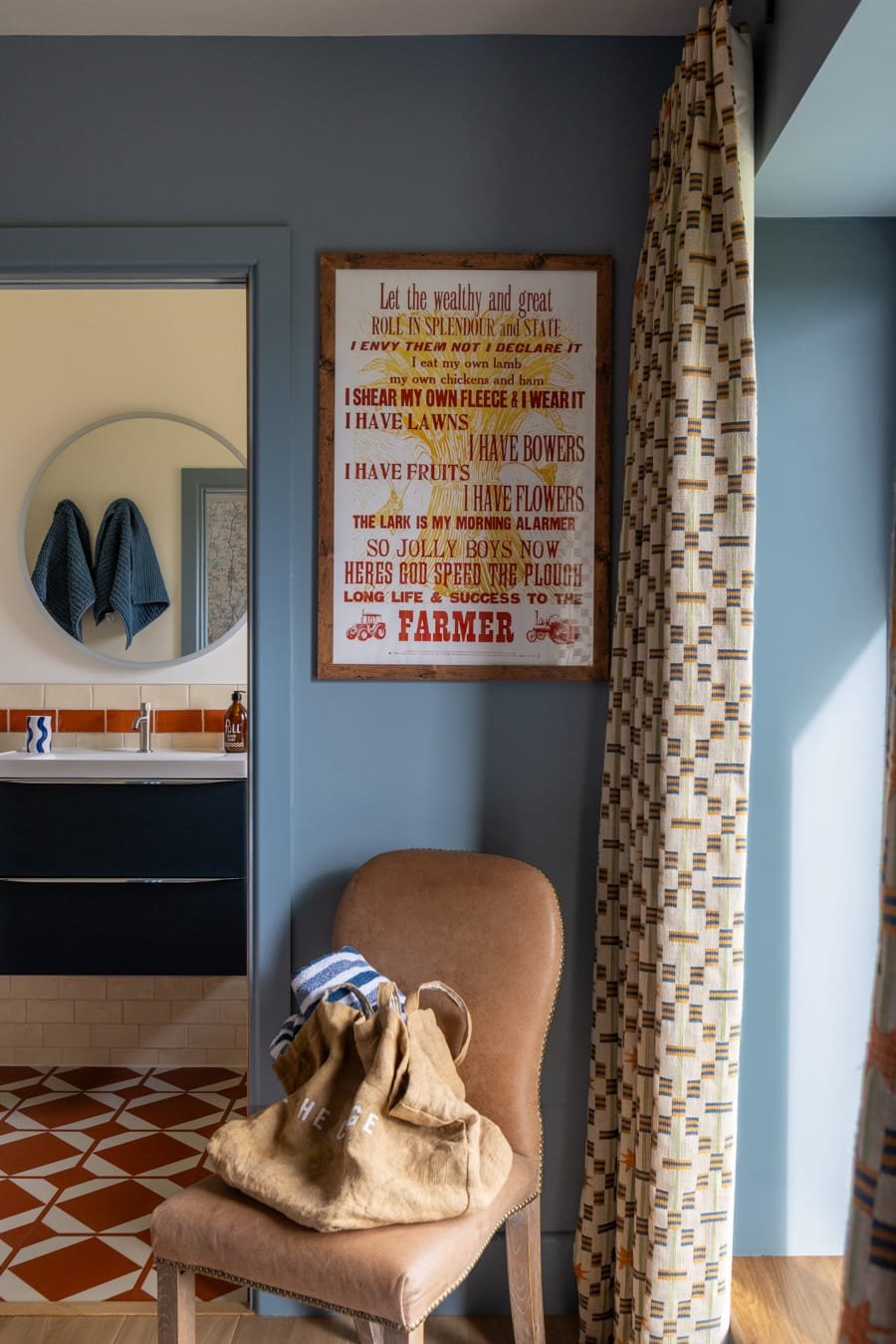 Interior of a room with a beige upholstered chair holding a tote bag and a striped cloth, a wall decorated with a framed quote, a curtain with a geometric pattern, a bathroom sink with two blue towels reflected in the mirror, and patterned brown and 