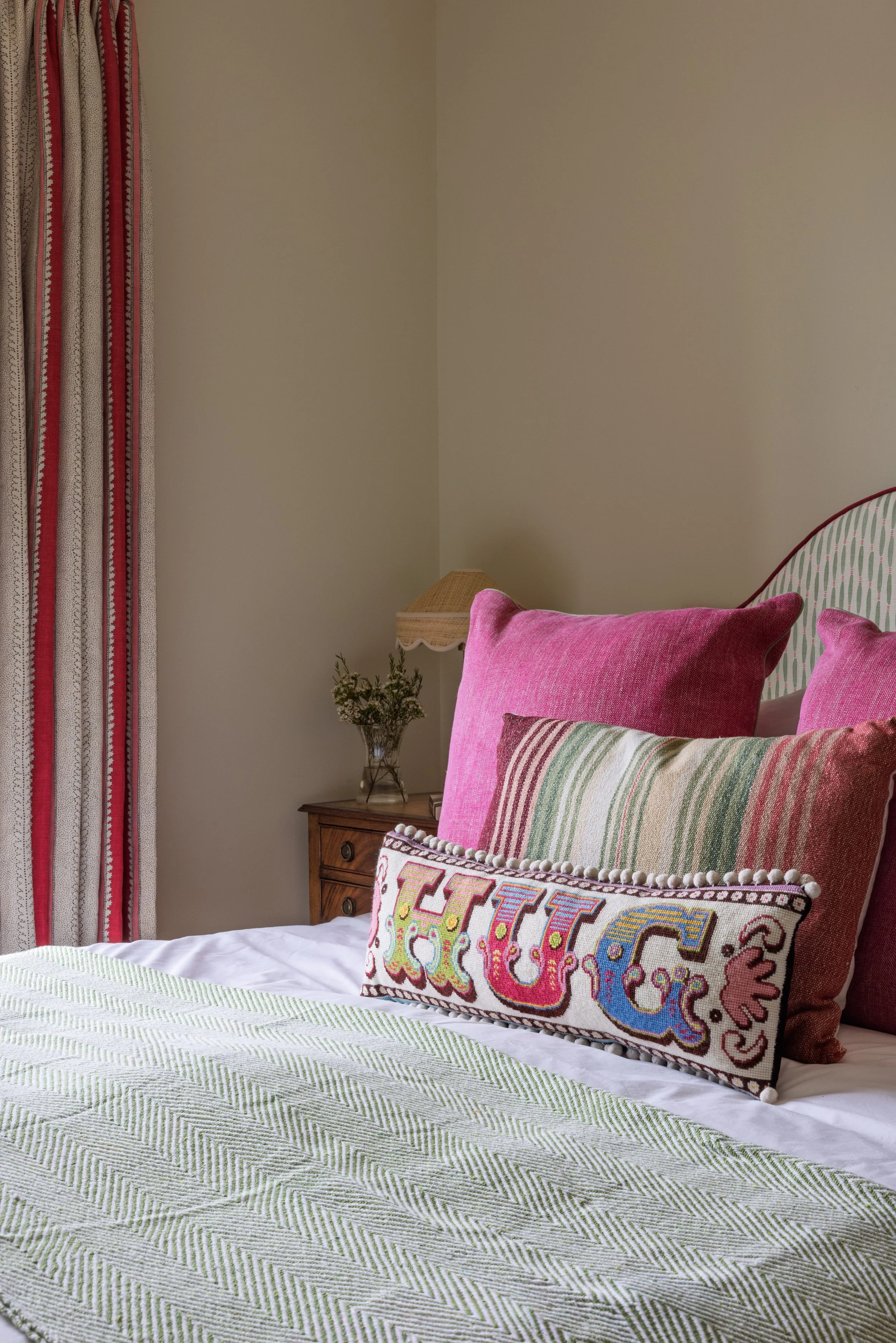 A bedroom with a bed covered with a white and green patterned bedspread, decorated with pink and striped pillows. To the left, a wooden nightstand with a small plant and a lampshade. There are red and white striped curtains on the window, and a beige