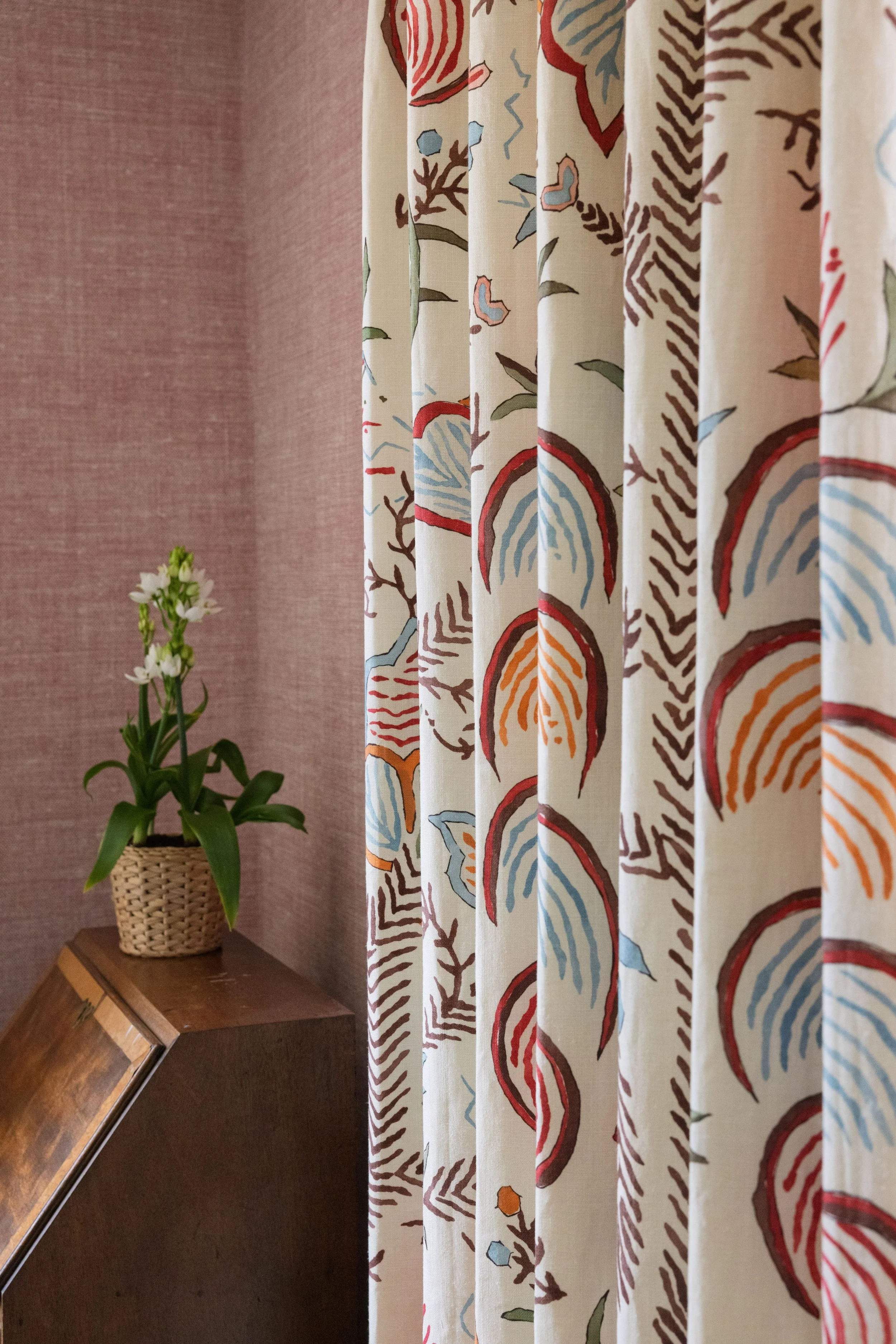 Close-up view of a floral-patterned curtain with circles, leaves, and abstract designs in red, orange, blue, and brown on a cream background. A potted plant with white flowers is on a wooden table next to a textured pink wall.