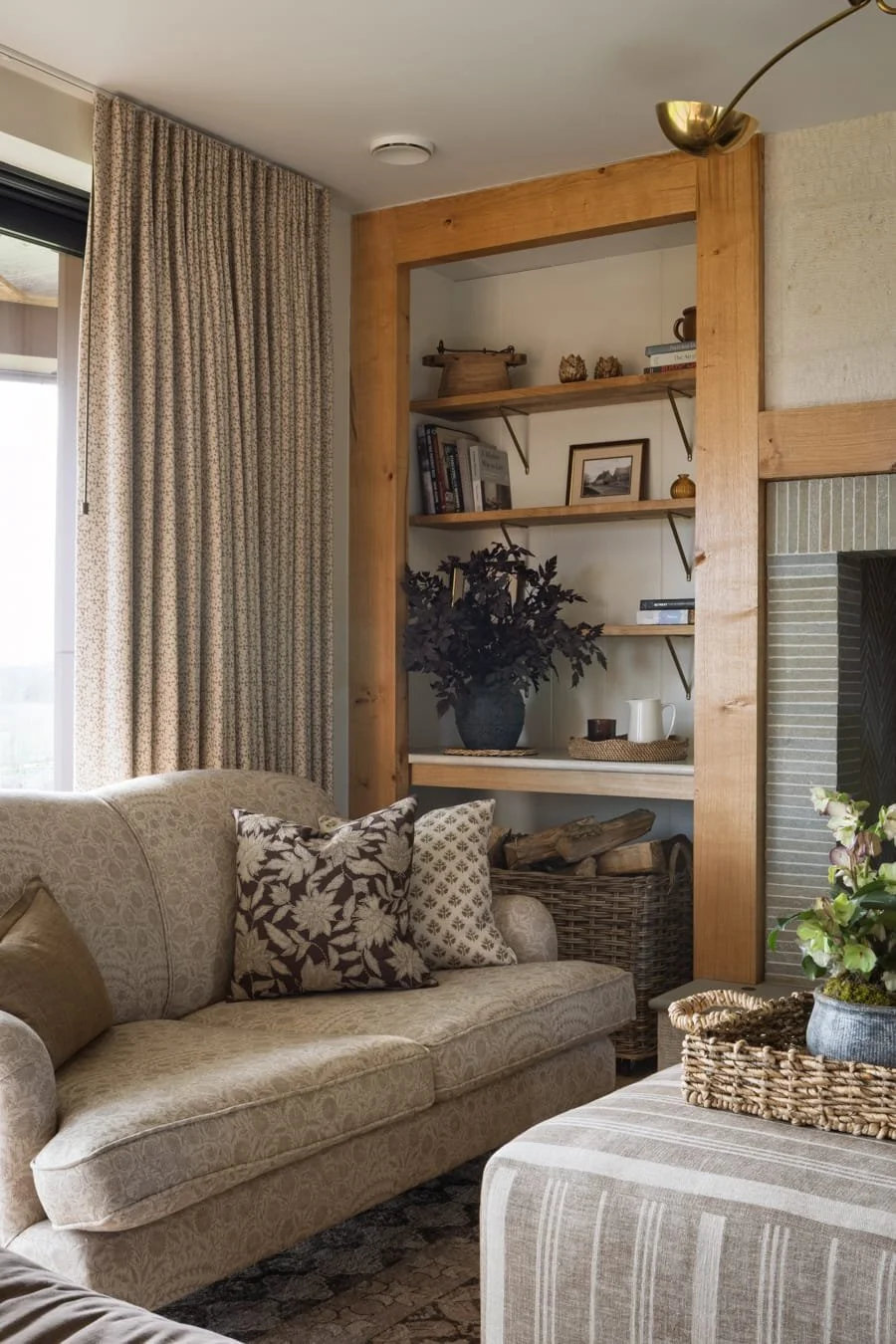 A cozy living room corner with a beige patterned sofa, decorative pillows, and a striped ottoman with a plant on top. A built-in wooden bookshelf with framed photos, books, and decorative objects is adjacent to a fireplace with a brick surround.