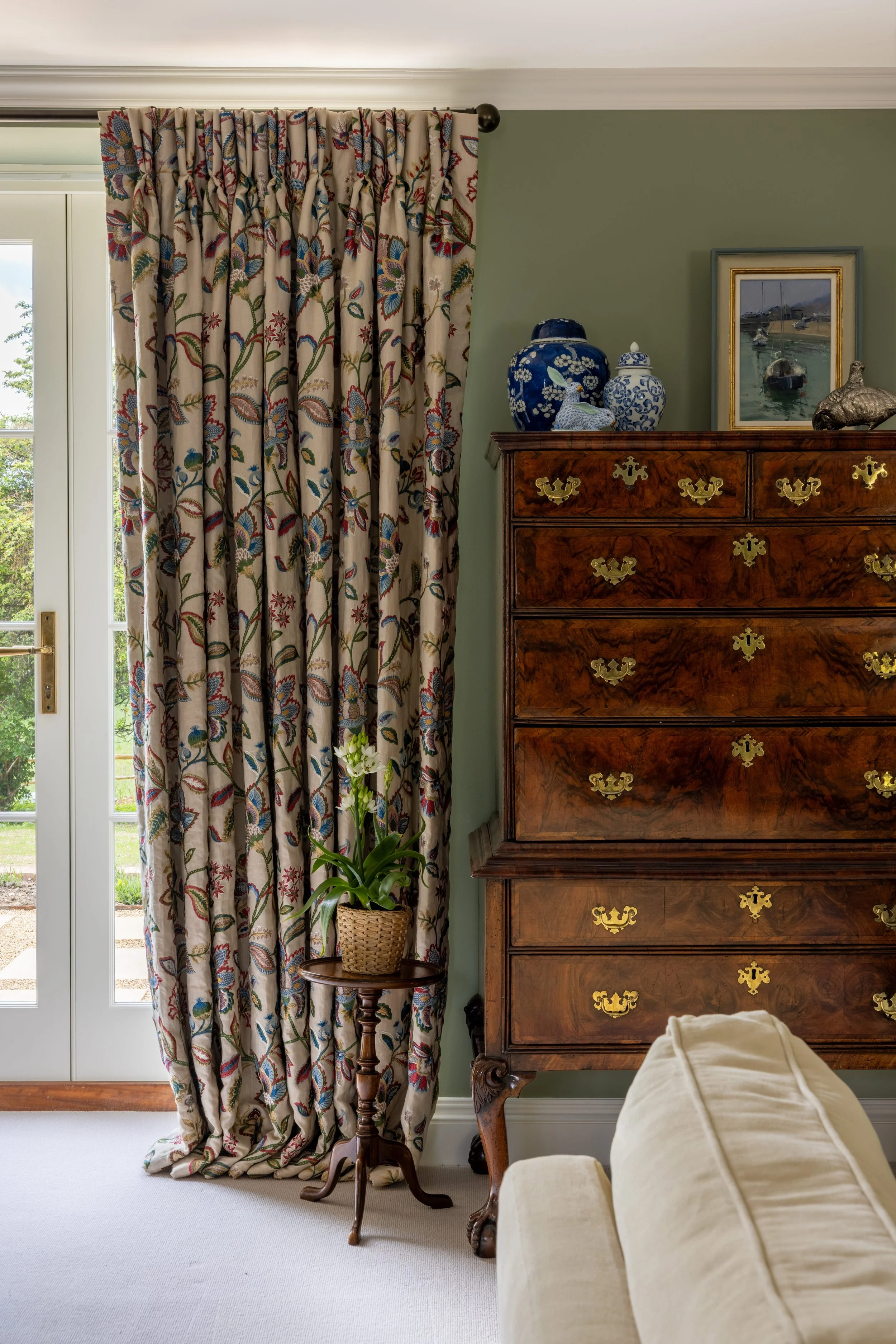 A living room corner with floral-patterned curtains, a potted plant on a small wooden table, an antique wooden dresser with brass handles, a framed painting of boats in a harbor, and a sofa in the foreground.