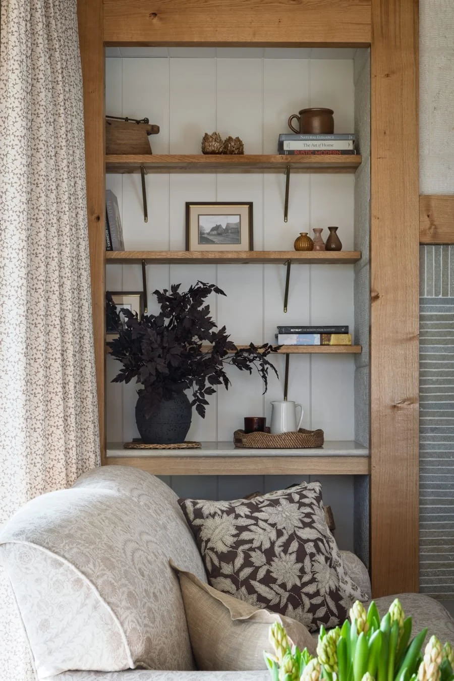 Close-up view of a living room with a built-in wooden shelf filled with decorative items, framed photos, books, a dark vase with purple foliage, and a beige and brown sofa with patterned cushions.