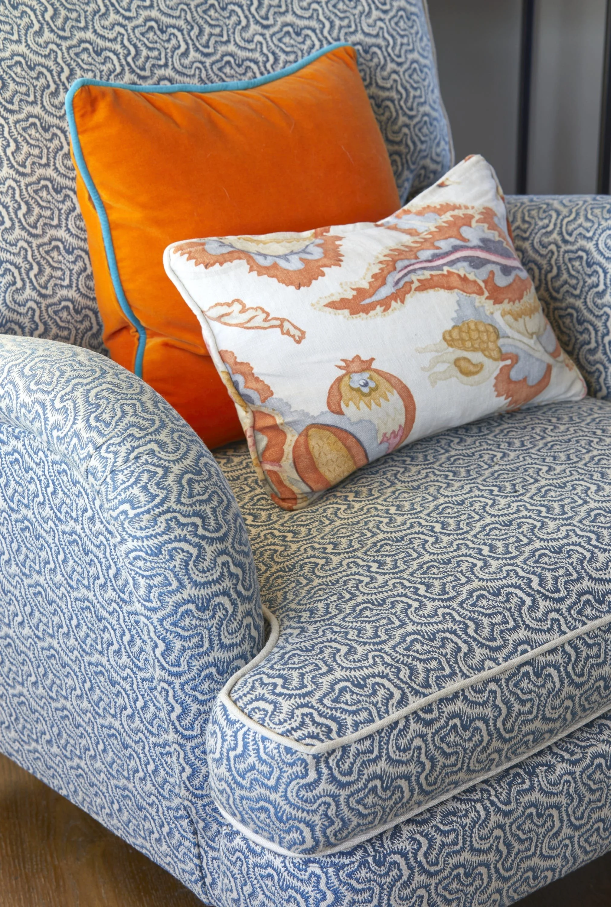 Close-up of a patterned blue and white upholstered armchair with two cushions: an orange cushion with a blue trim and a decorative cream-colored cushion with an orange and blue floral pattern.