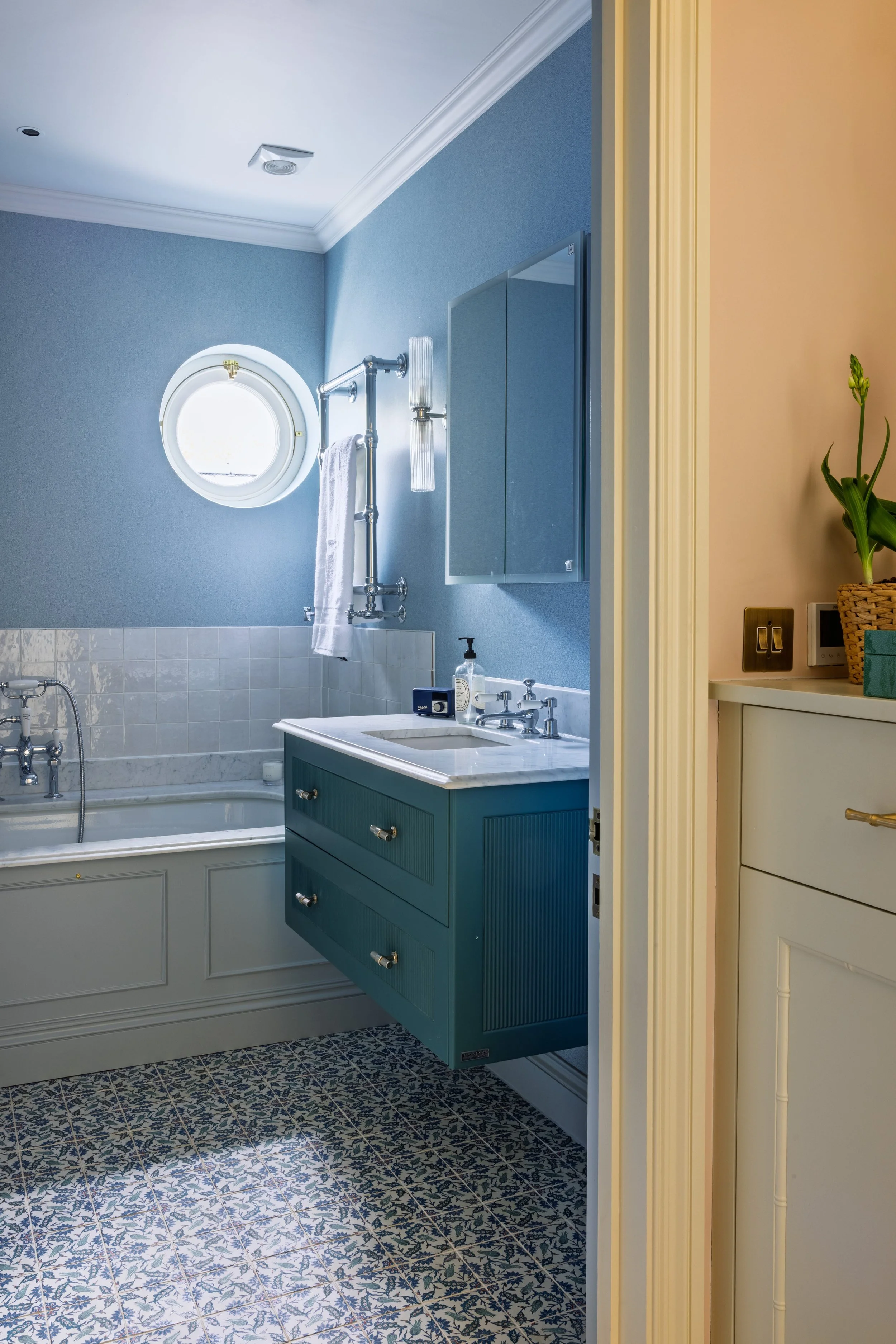 Reflected image of blue bathroom featuring a small vanity with a blue cabinet, a mirror, a window with a porthole design, and a pipe with a towel attached. Part of a plant and a beige wall are visible outside the bathroom.