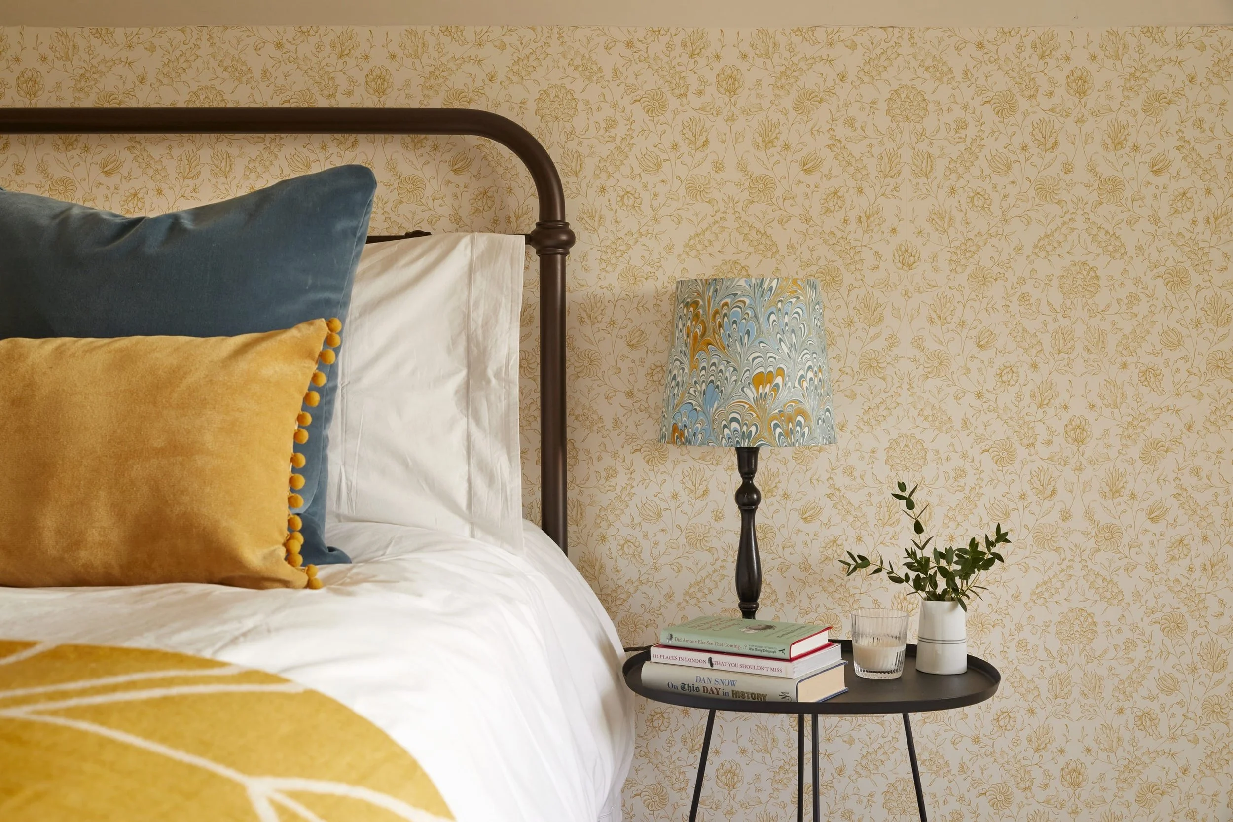 A cozy bedroom corner featuring a bed with blue and yellow pillows, a black nightstand holding books, a lamp with a swirling blue, gold, and white lampshade, and a plant in a white pot, all against a patterned vintage wallpaper.