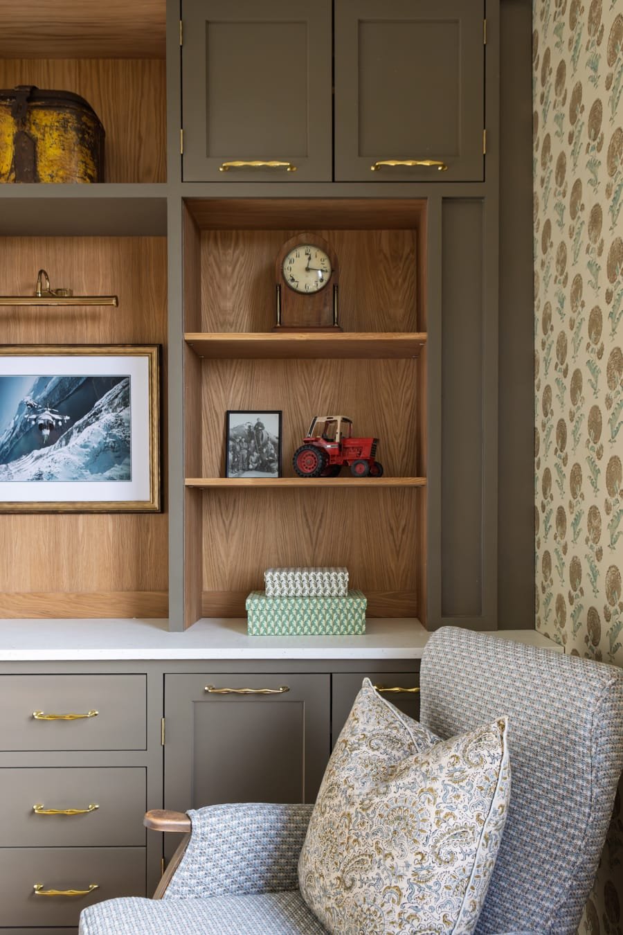 A built-in wooden shelf with gray cabinets and gold handles, decorative items including a vintage clock, framed photos, a toy tractor, and a box of tissues, next to a patterned armchair with a pillow, and a wall with floral wallpaper.