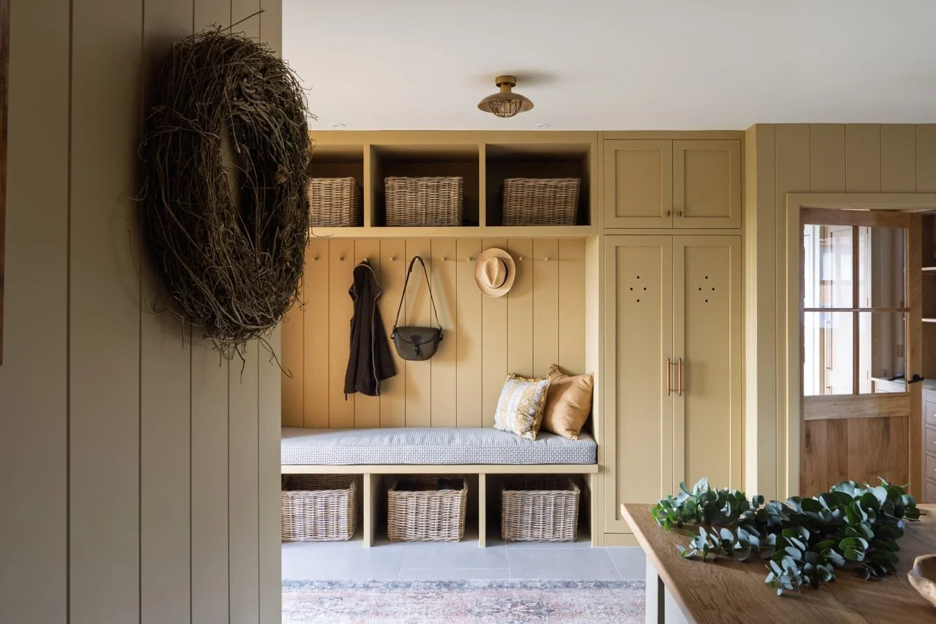 Entryway with built-in bench, storage baskets, hooks with hats and bags, and beige cabinetry in a rustic style.