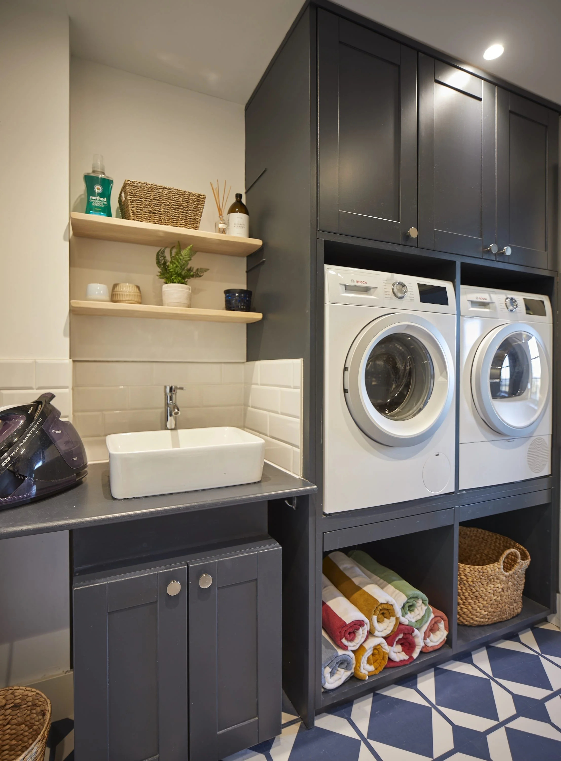 Laundry room with dark gray cabinetry, two front-loading washing machines, open shelving with towels and baskets, a small sink, and decorative items on the shelving.