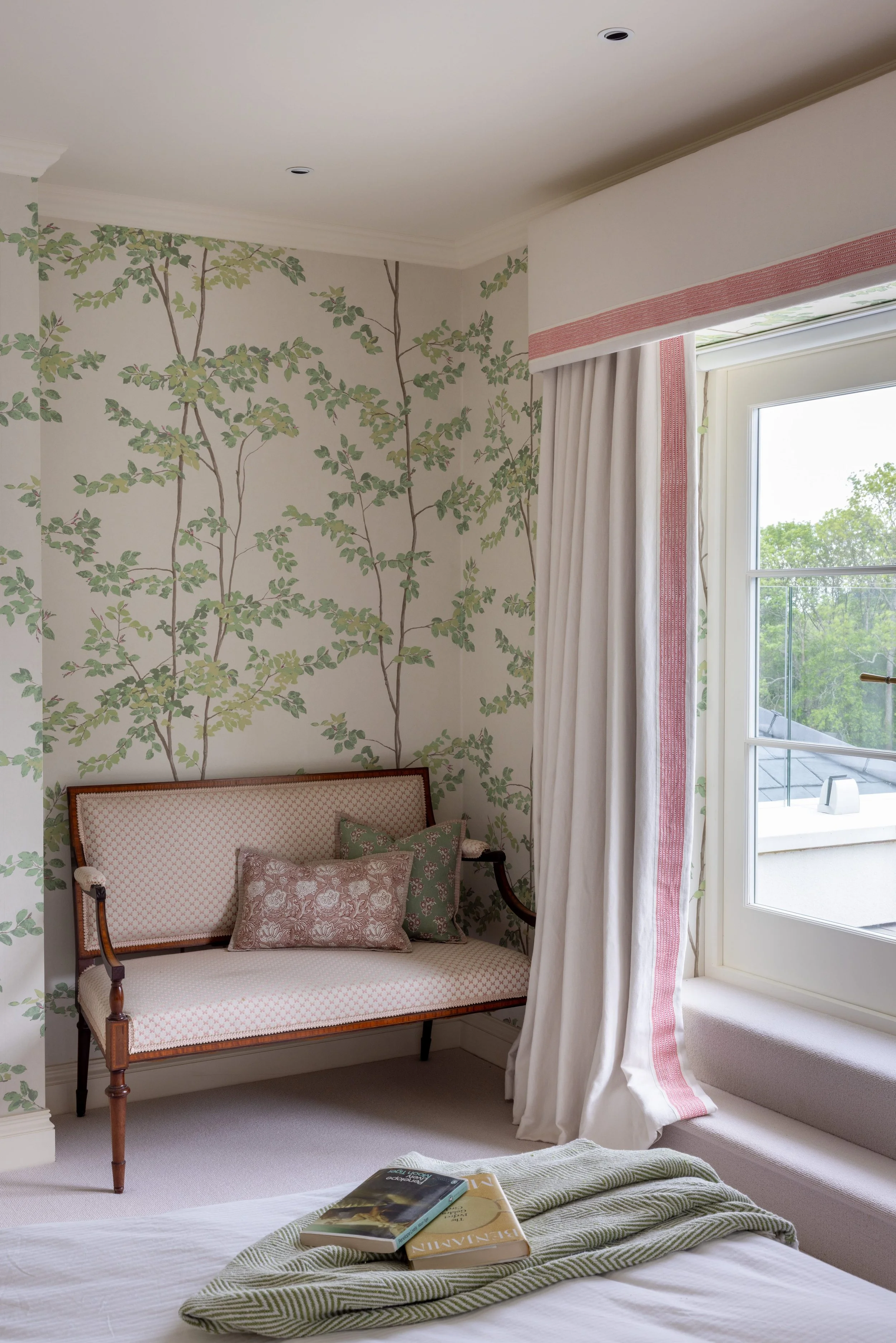 A cozy bedroom corner with a vintage wooden settee decorated with patterned cushions, an wallpaper with leafy green branch pattern, an open window with white curtains, and a bed with books on a green and white striped blanket.