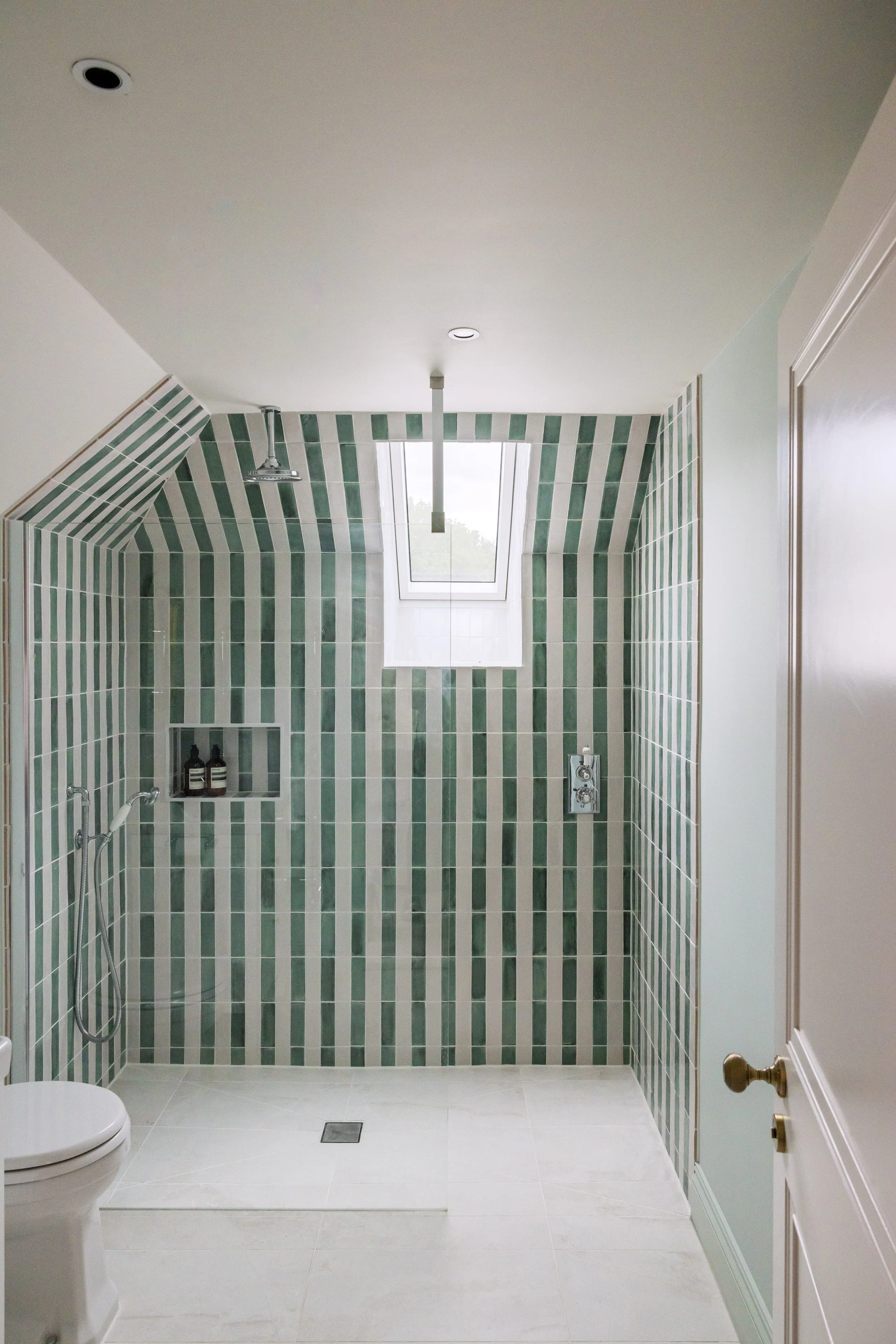 Bathroom with green and white striped tiles, a skylight window, a shower with a hand-held showerhead, a recessed shelf with toiletries, and a white toilet. Off-white walls and door with a bronze doorknob.