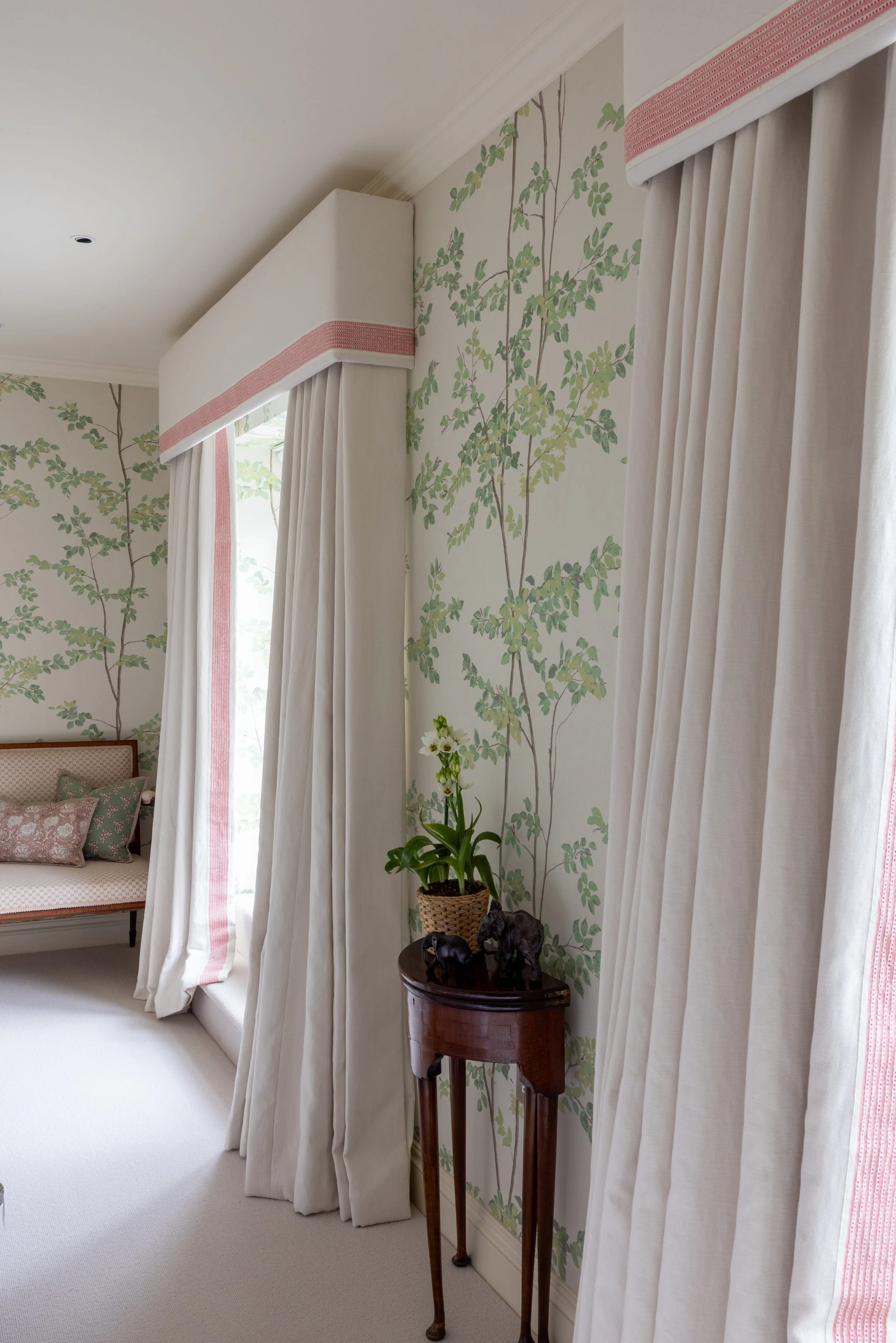 A corner of a room with white curtains, a plant on a wooden stand, and a sofa with patterned pillows.