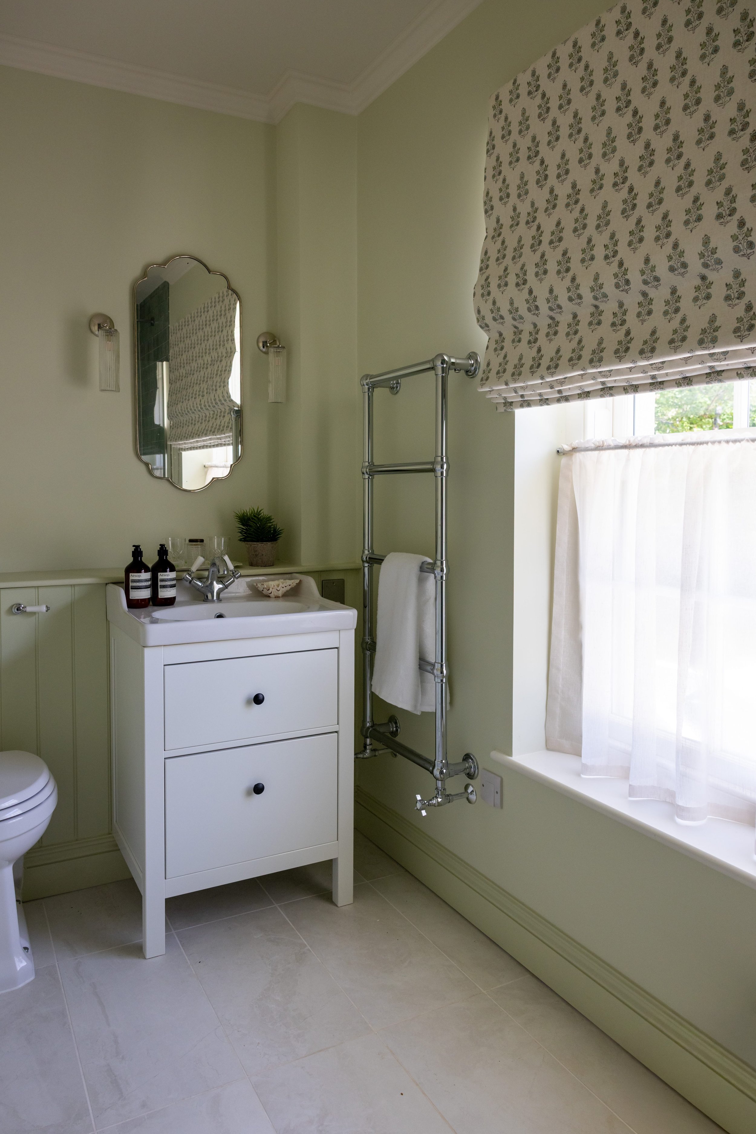 A bathroom with a white vanity, small mirror, potted plant, two soap bottles, and a wall-mounted towel rack. The window has a floral Roman shade and a sheer curtain, with light green painted walls and tile flooring.
