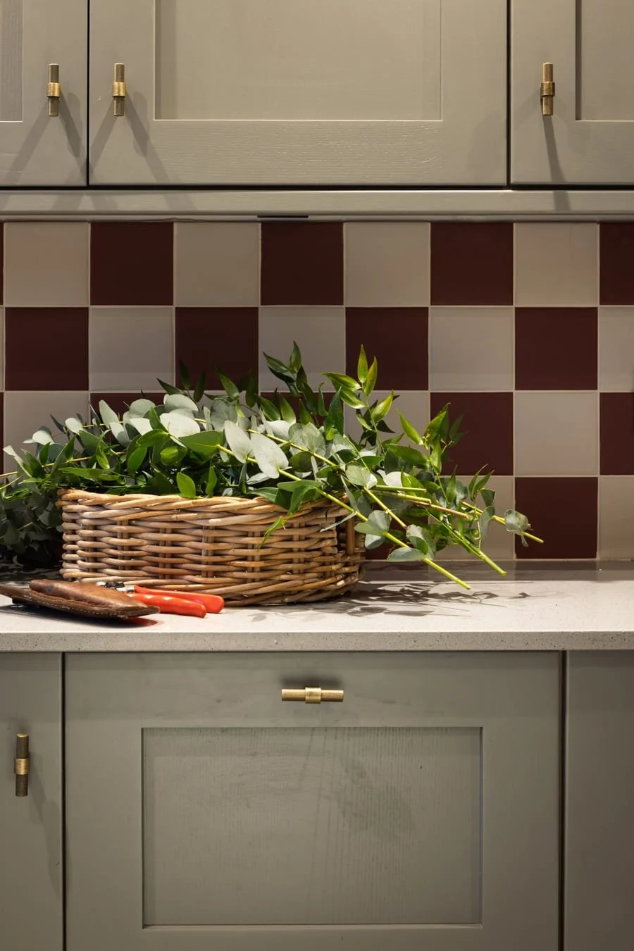 A kitchen countertop with a wicker basket filled with green leafy branches, garden pruning tools, and a tiled backsplash with a checkered pattern of red and white tiles.