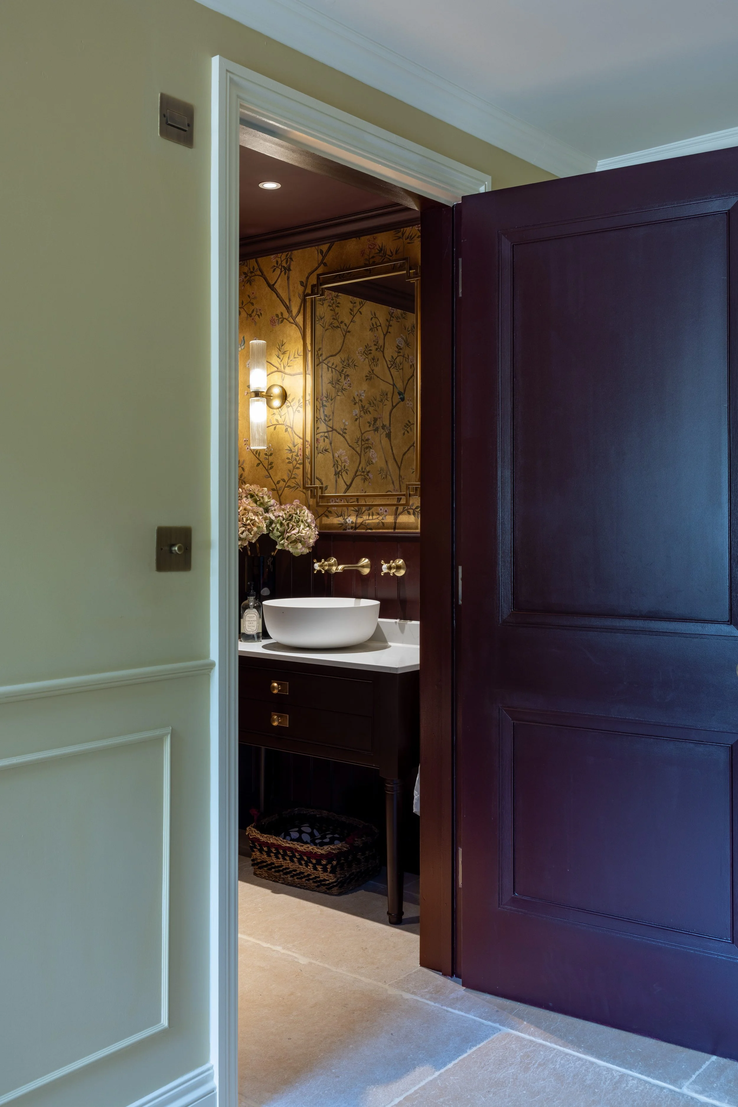 Partially opened dark wooden door reveals a bathroom with wallpaper featuring a tree and bird pattern, a wall-mounted light, a white vessel sink, a wicker wastebasket, and beige tile flooring.