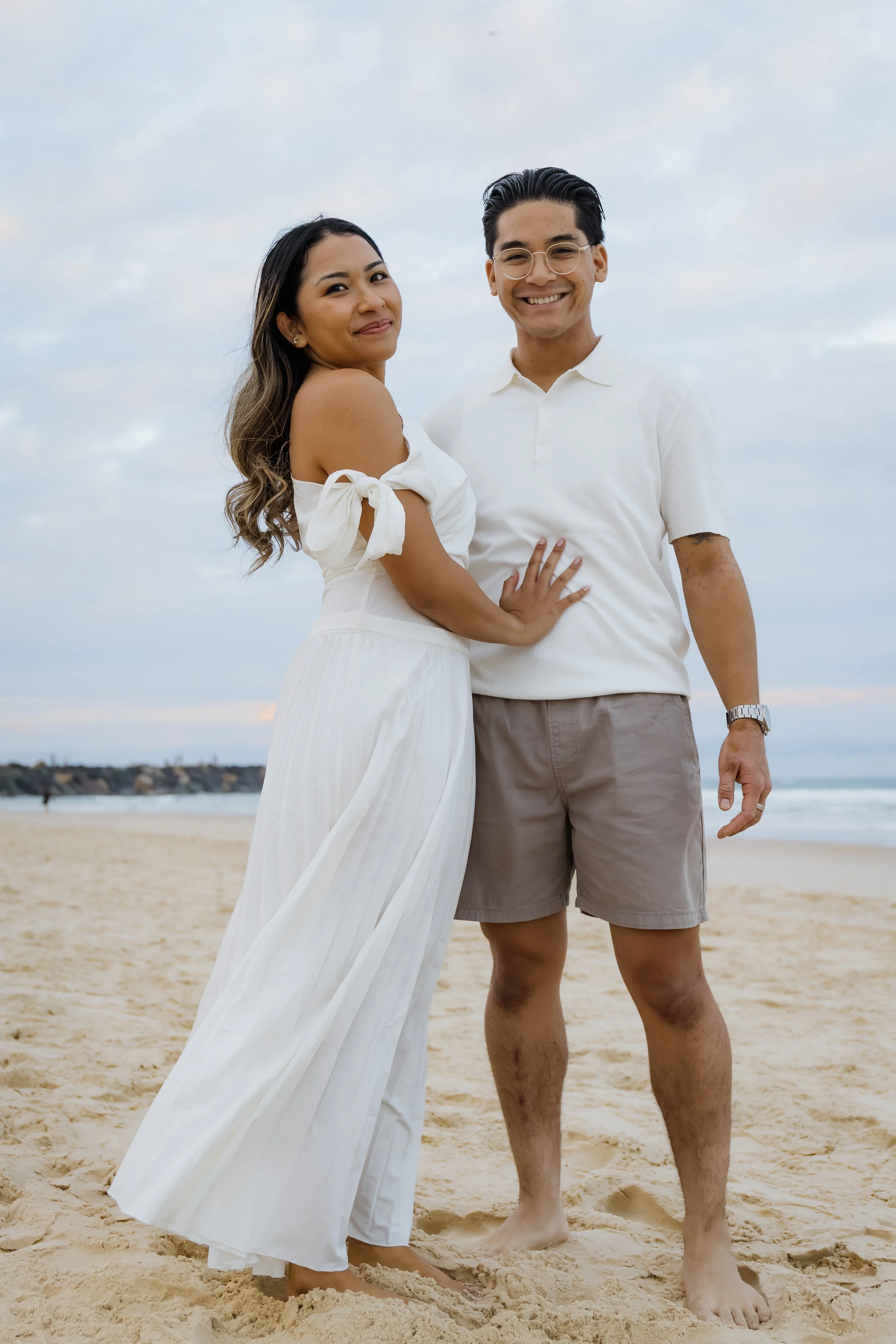 A smiling couple standing on a sandy beach with a cloudy sky behind them. The woman is wearing a white off-shoulder dress and the man is wearing a white polo shirt and beige shorts.
