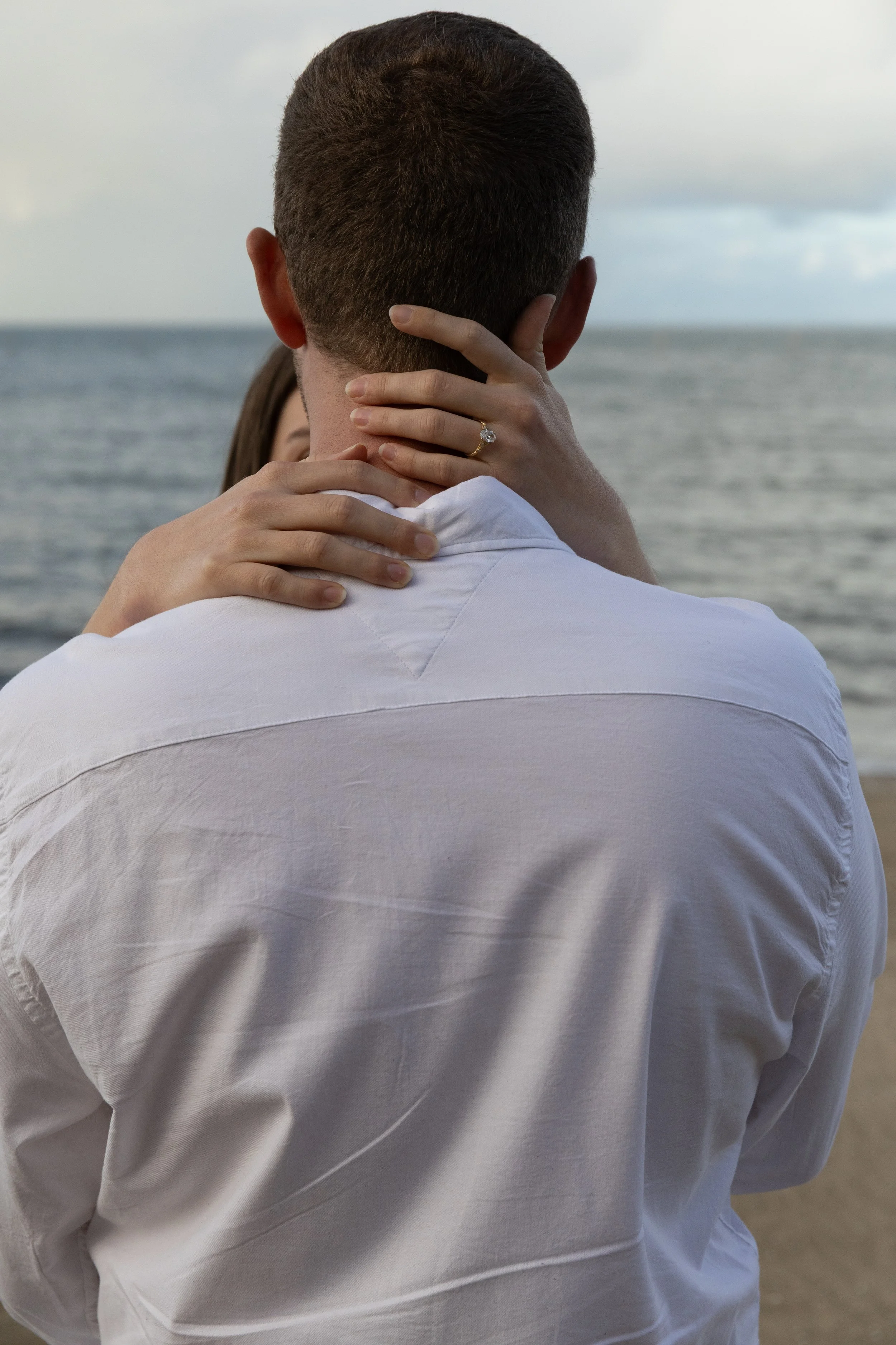 A man with short dark hair, wearing a white shirt, facing away from the camera, with a woman’s hand around his neck, showing an engagement ring. They are at the beach with the ocean and sky in the background.