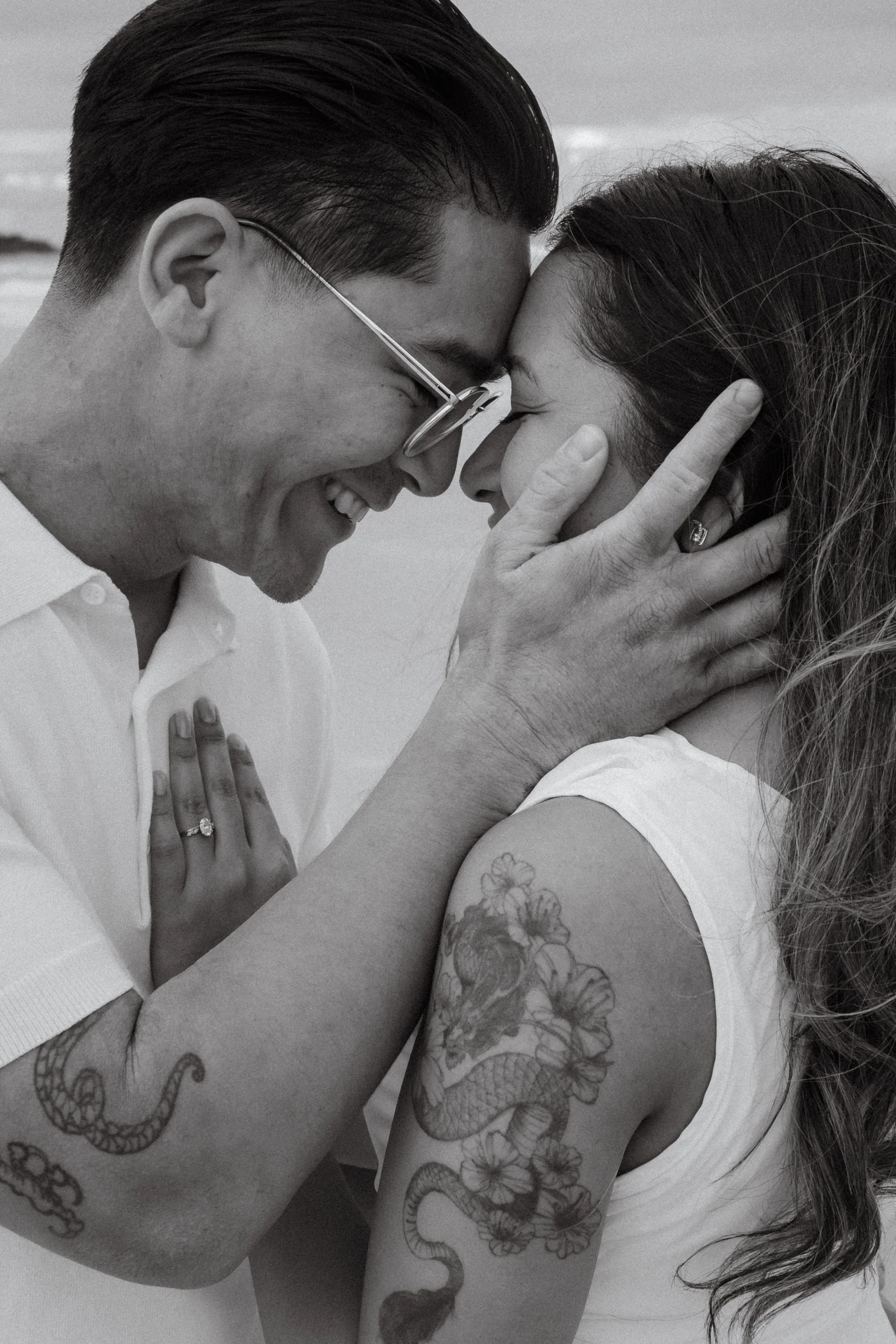 A man and woman touching foreheads with eyes closed, smiling, holding each other's faces, and embracing, with visible tattoos on their arms.