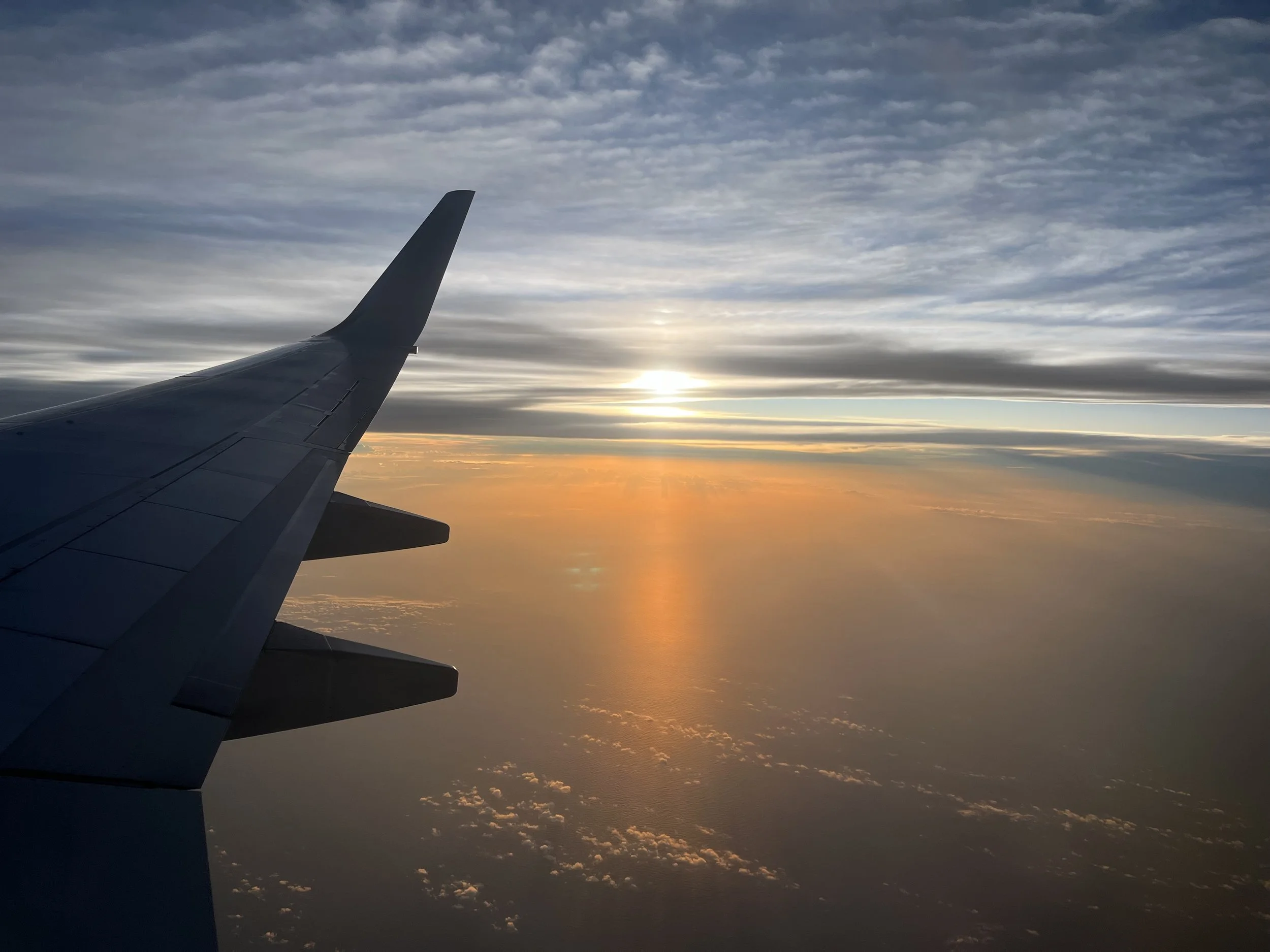 View from an airplane window showing the wing and a sunset sky with clouds and a colorful horizon.