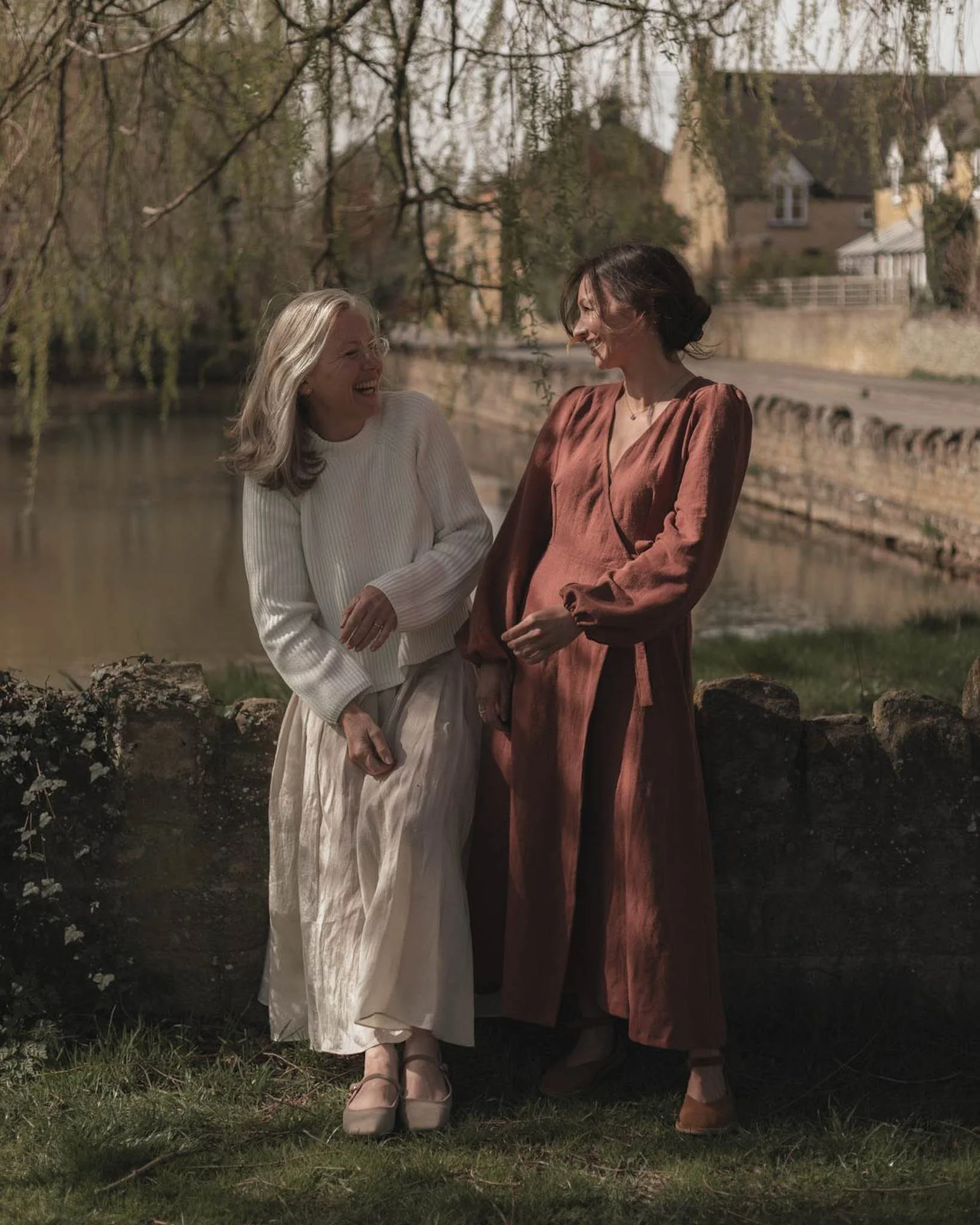 Two women standing outdoors near a river, laughing and talking, with trees and houses in the background.